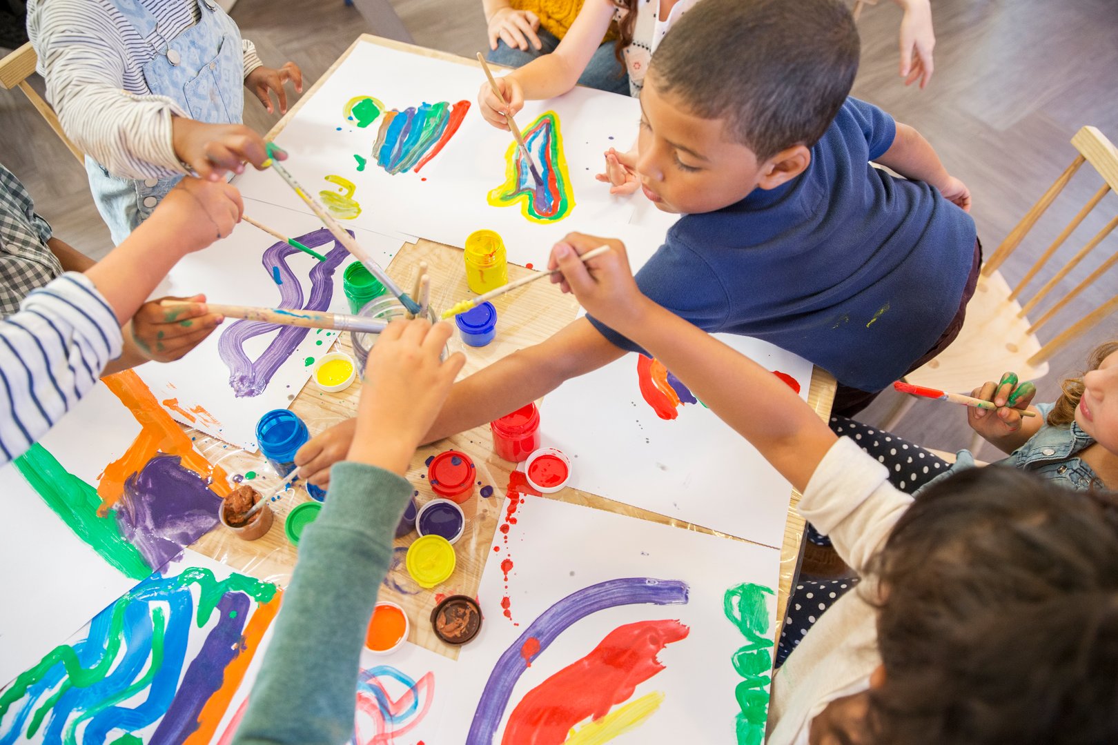 Children gathered around a table, painting colorful designs on paper with brushes and paint jars.