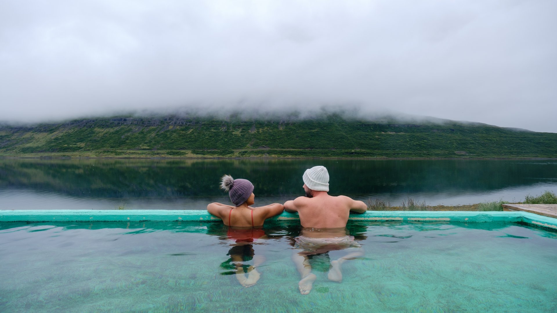 A couple unwinds in a geothermal hot spring surrounded by serene Icelandic nature. Clouds hover over the lush green hills, creating a calm atmosphere perfect for relaxation Westfjords Iceland