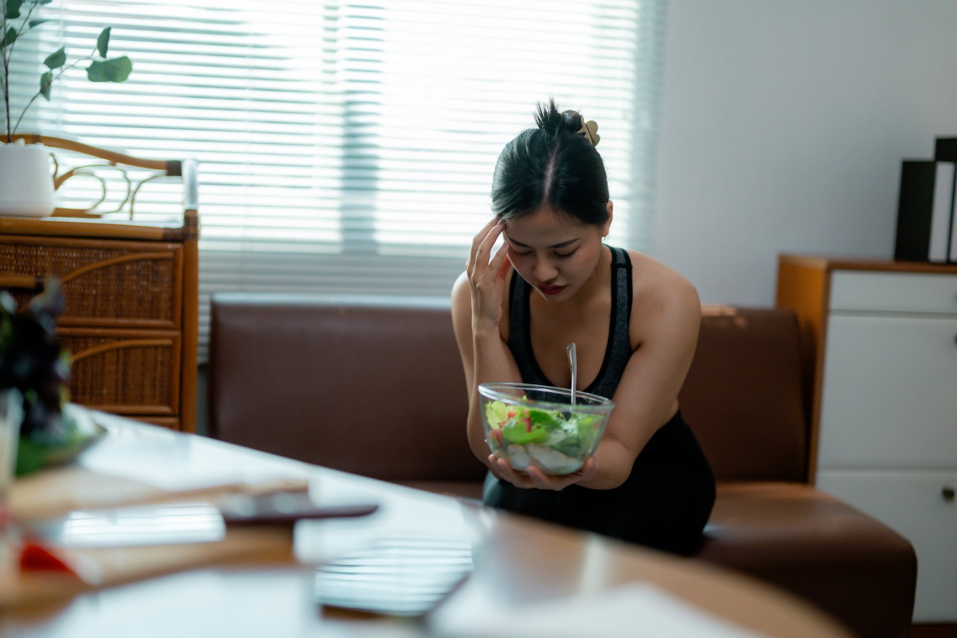 Young asian woman suffering from anorexia looking at a salad bowl while sitting on the sofa in the living room at home, anorexia and bulimia nervosa concept