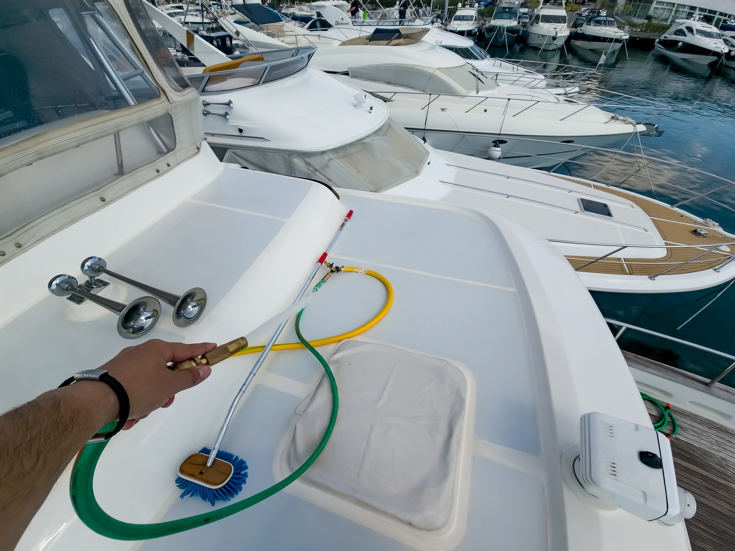 Close-up perspective of marine maintenance professional using specialized cleaning brush and hose on white yacht deck, with luxury vessels docked in background