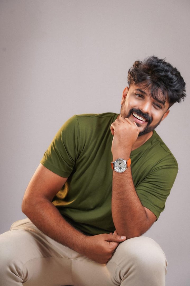 A man in a green shirt and beige pants smiles, resting his chin on his hand. He wears a watch and sits against a neutral background.