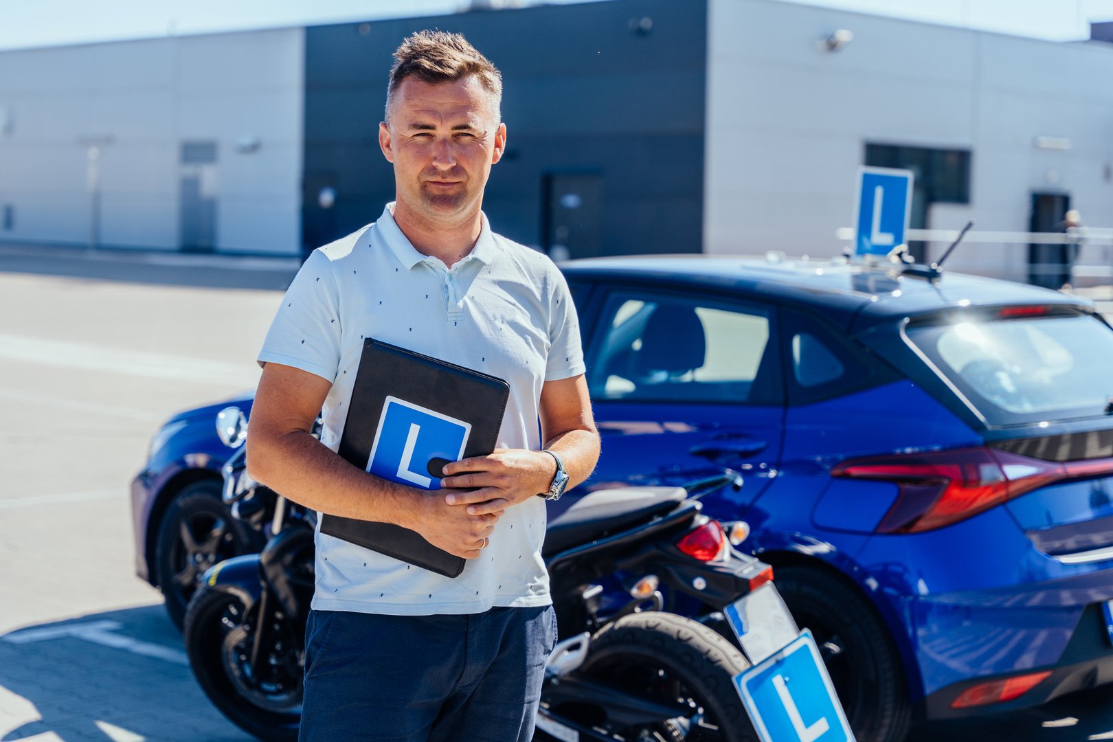 Portrait of middle-aged male driving instructor stands beside motorcycle and training car with clipboard. Motorcycle driving school.