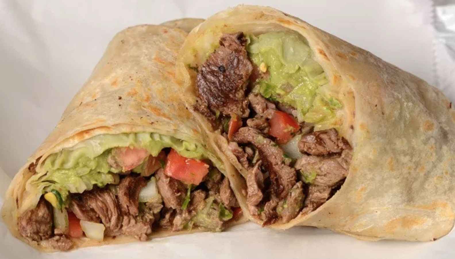 A beef burrito cut in half, filled with grilled steak, lettuce, tomatoes, and guacamole, wrapped in a flour tortilla.