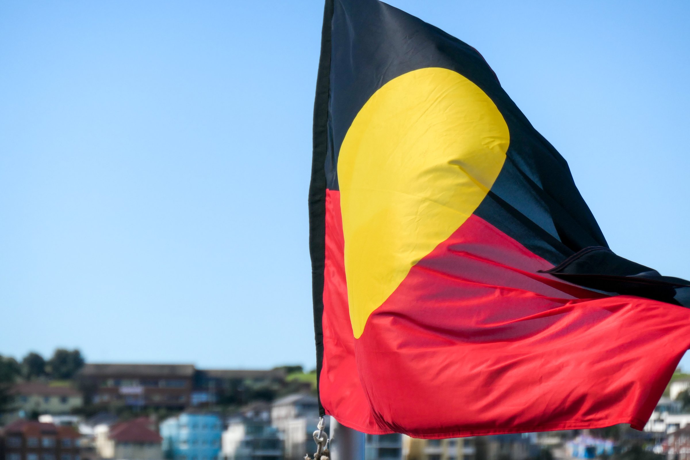 The Australian Aboriginal flag flies in the wind with the background of Bondi Beach, Sydney, on a winter afternoon in June 2022.  The flag is being blown in the wind and yellow circle appears in the shape of a teardrop.