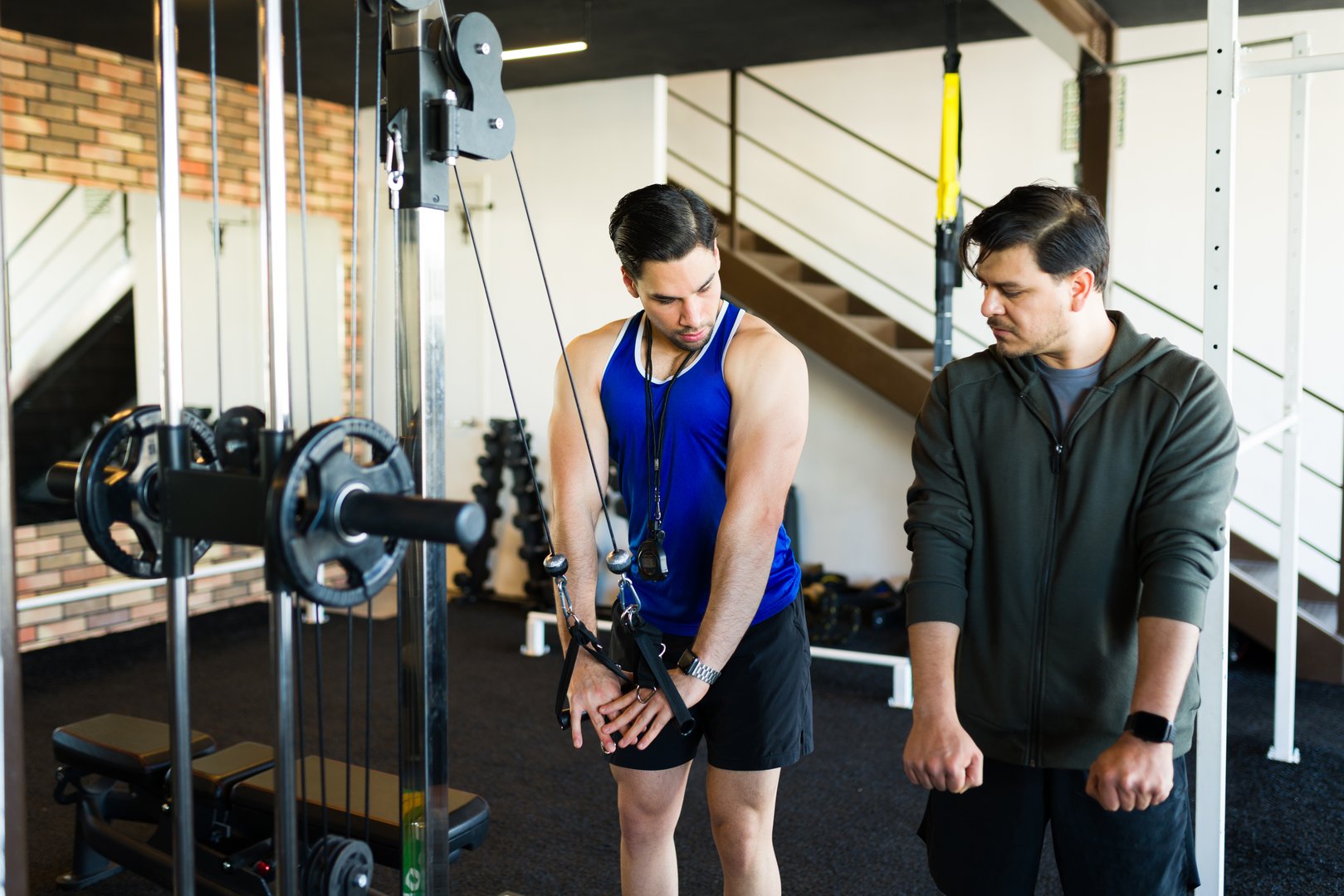 Hispanic man working out using cable crossover machine with the help of his personal fitness coach at the gym