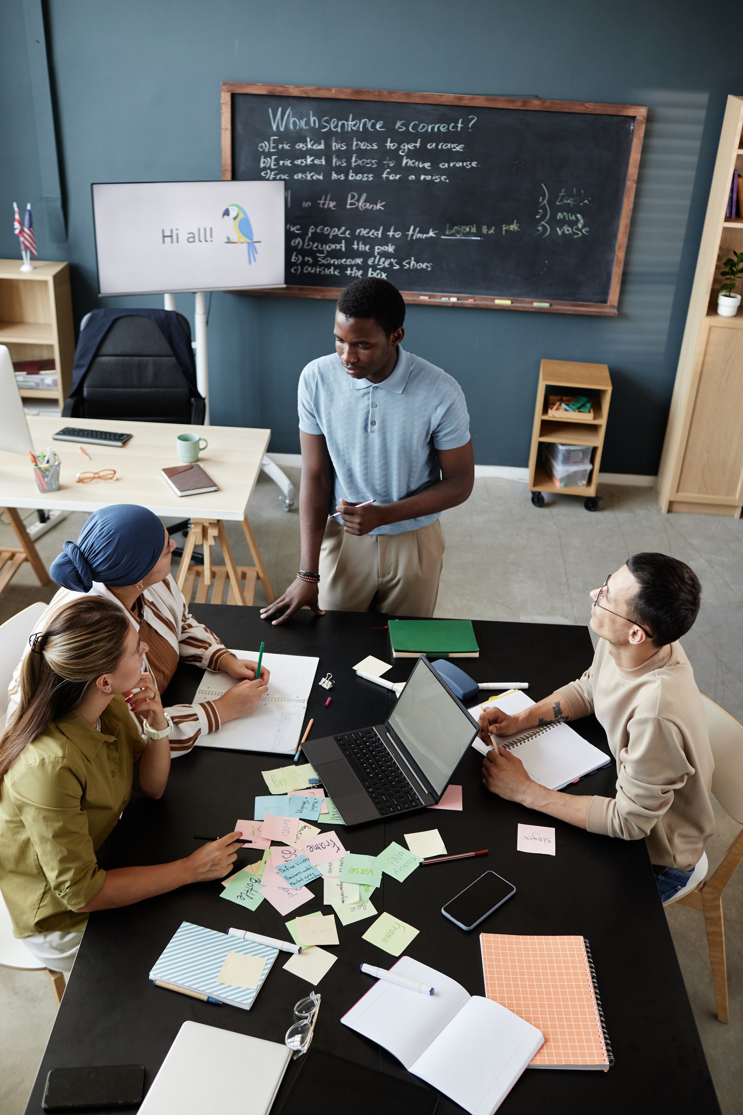 Vertical shot of young African American teacher standing at desk talking to students explaining task, during speaking club session in English language school for adult learners