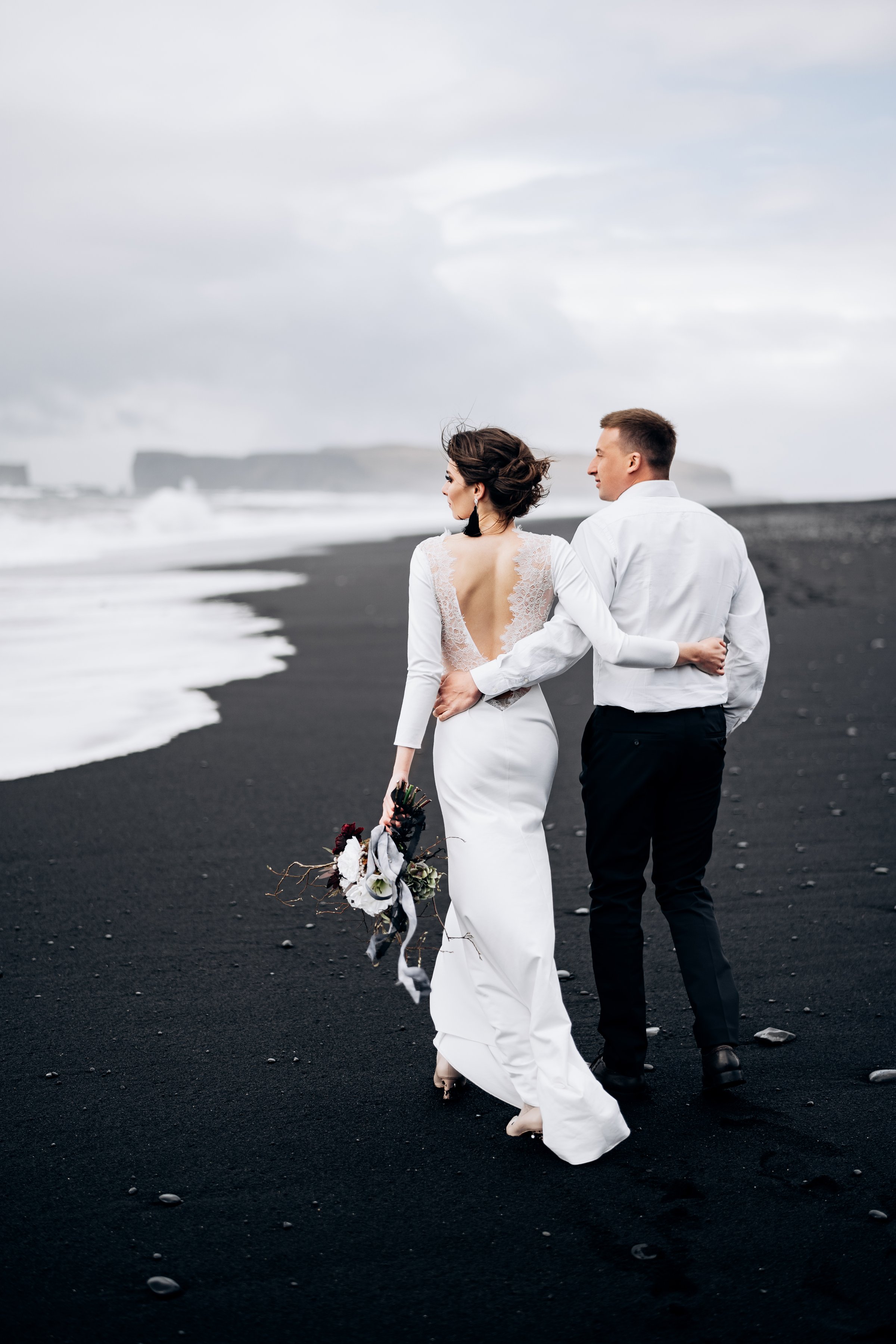Wedding couple walks on black Beach Vic. A sandy beach with black sand on the shores of the Atlantic Ocean. Huge frothy waves. The groom hugs the bride.