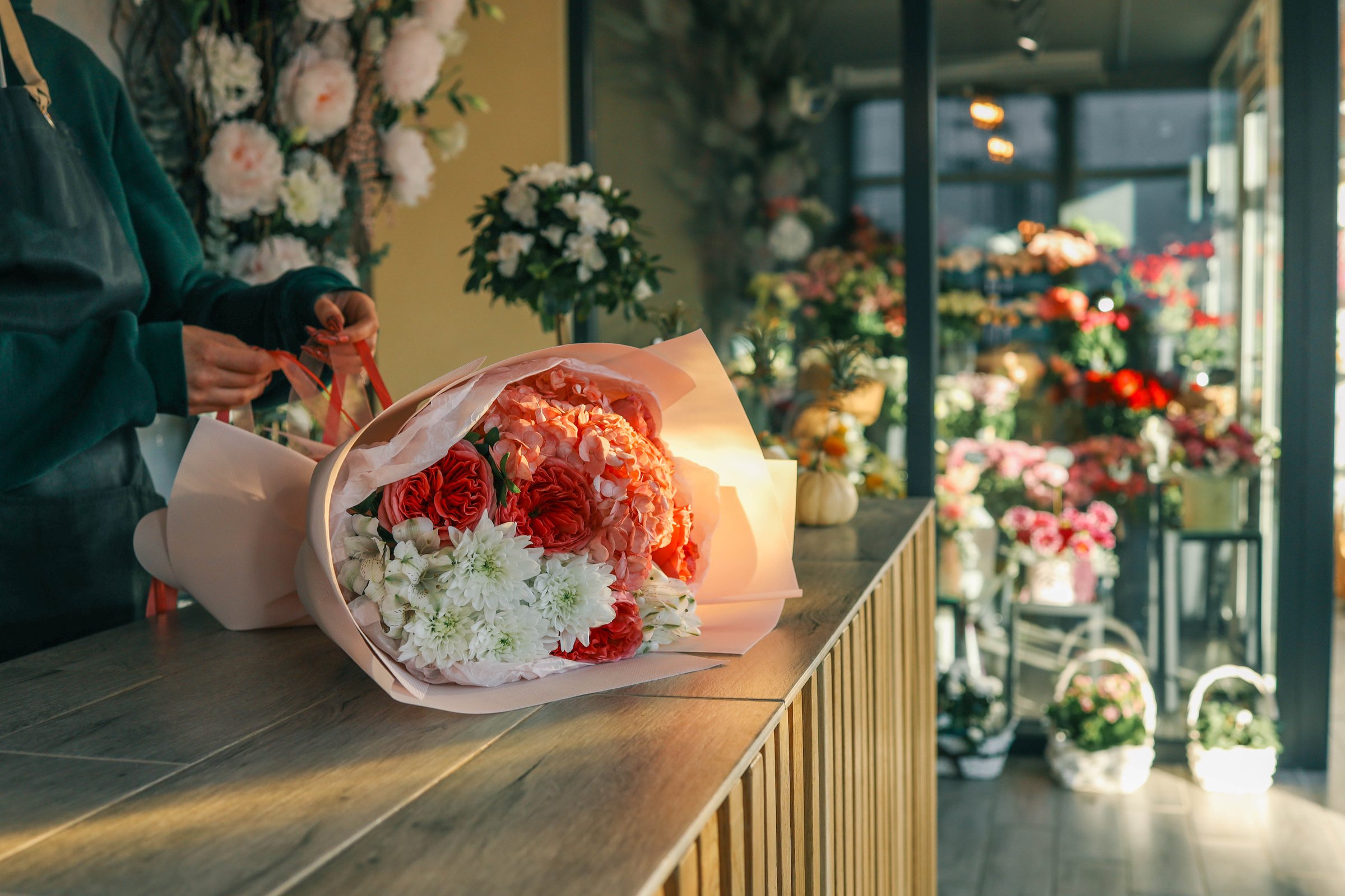 Close-up of a luxurious bouquet featuring vibrant red roses, delicate hydrangeas, and white alstroemerias, elegantly arranged and wrapped in soft pastel paper on a wooden counter.