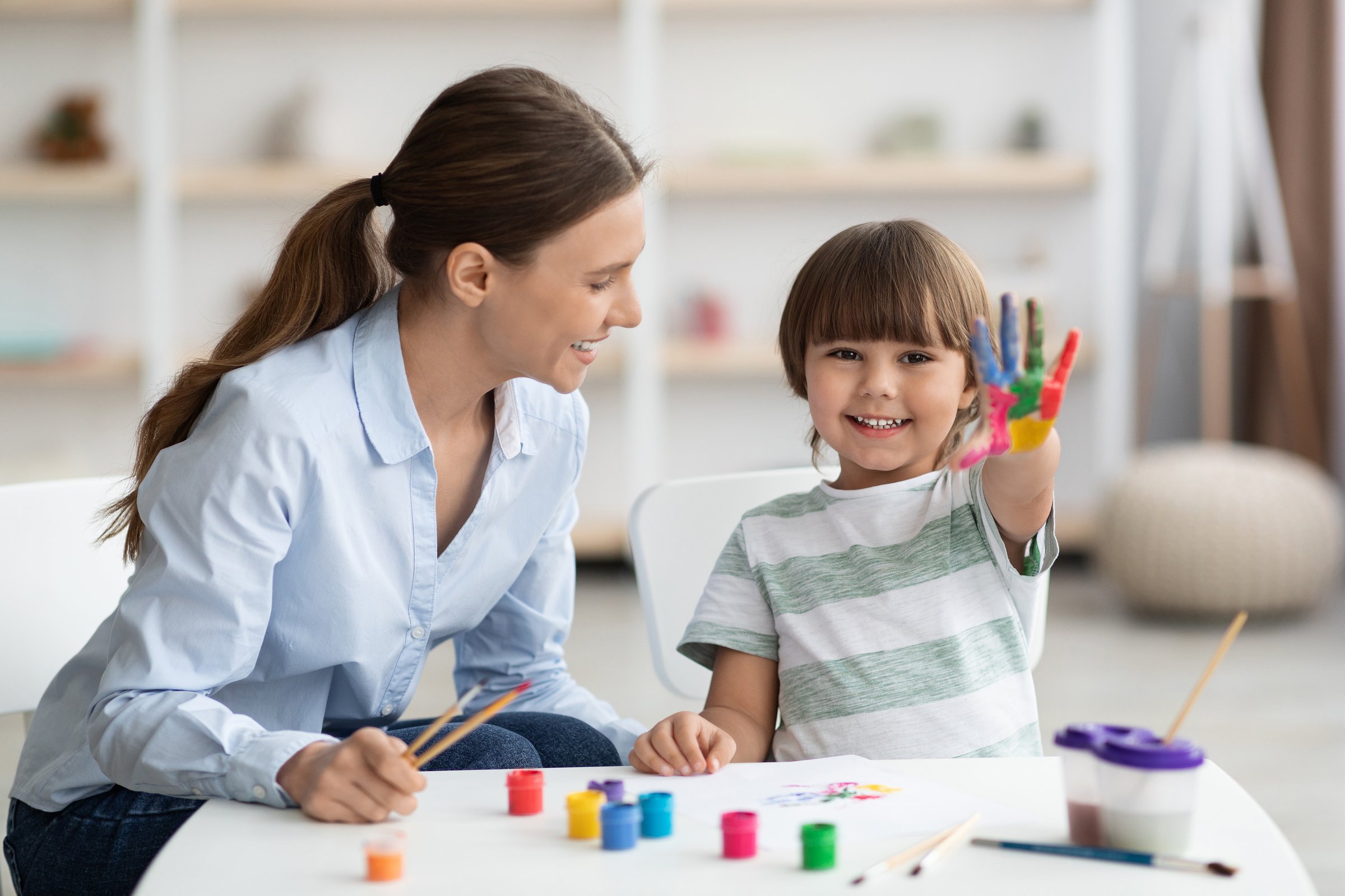Learning and education of kids. Happy little boy showing colorful palm, smiling to camera, sitting with professional psychologist during art therapy at office