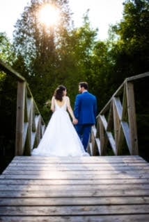 Bride and groom holding hands, walking on a wooden bridge with trees and sunlight in the background.