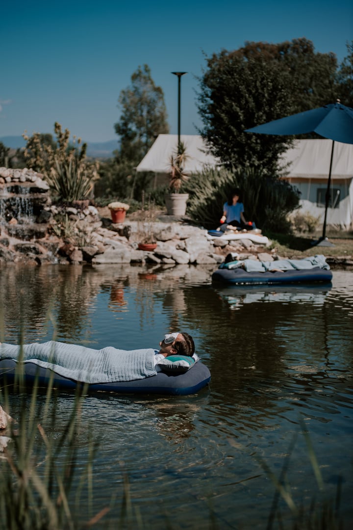 Person floating on an inflatable in a pond, another doing yoga nearby. Tents and greenery in background.