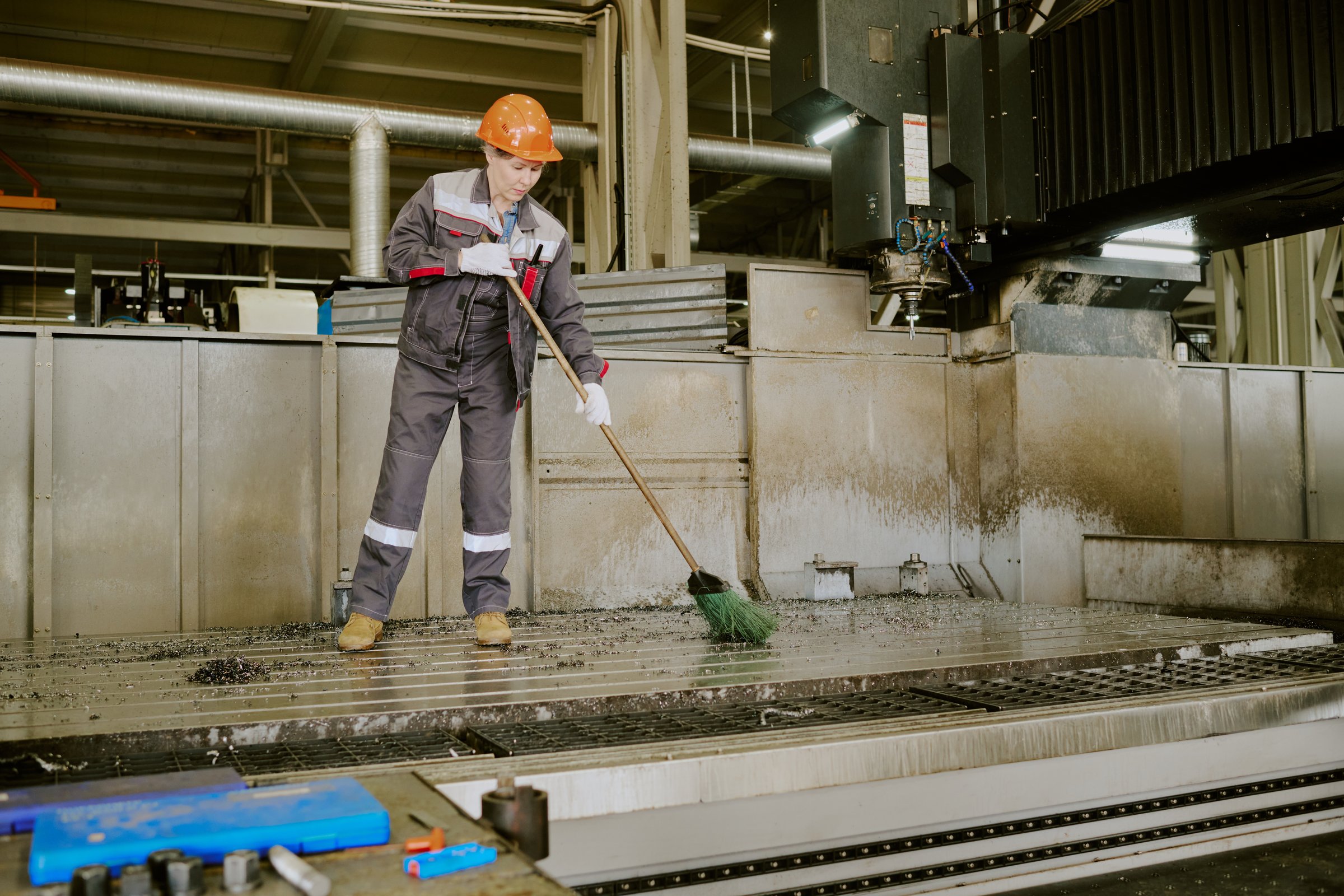 Middle aged Caucasian woman wearing protective helmet sweeping industrial workshop floor with broom, standing near large machinery, performing maintenance task in manufacturing facility