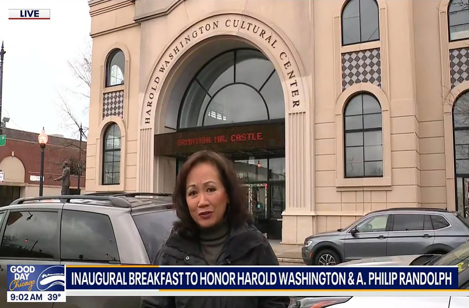 A news reporter stands outside the Harold Washington Cultural Center, with signage and a clock showing 9:02 AM.