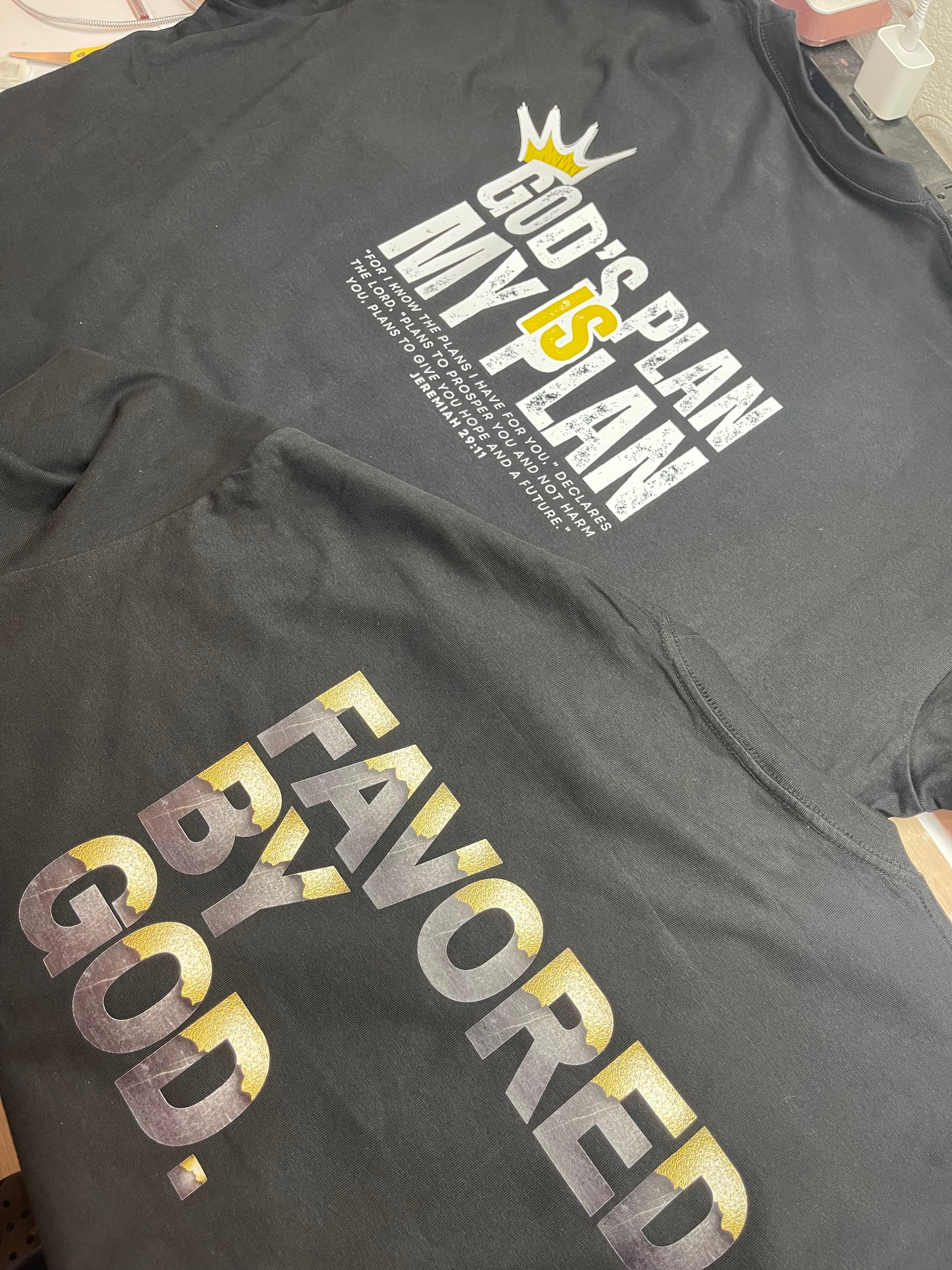 Two black T-shirts with religious messages: Gods Plan is My Plan and Favored by God, featuring bold, decorative text.