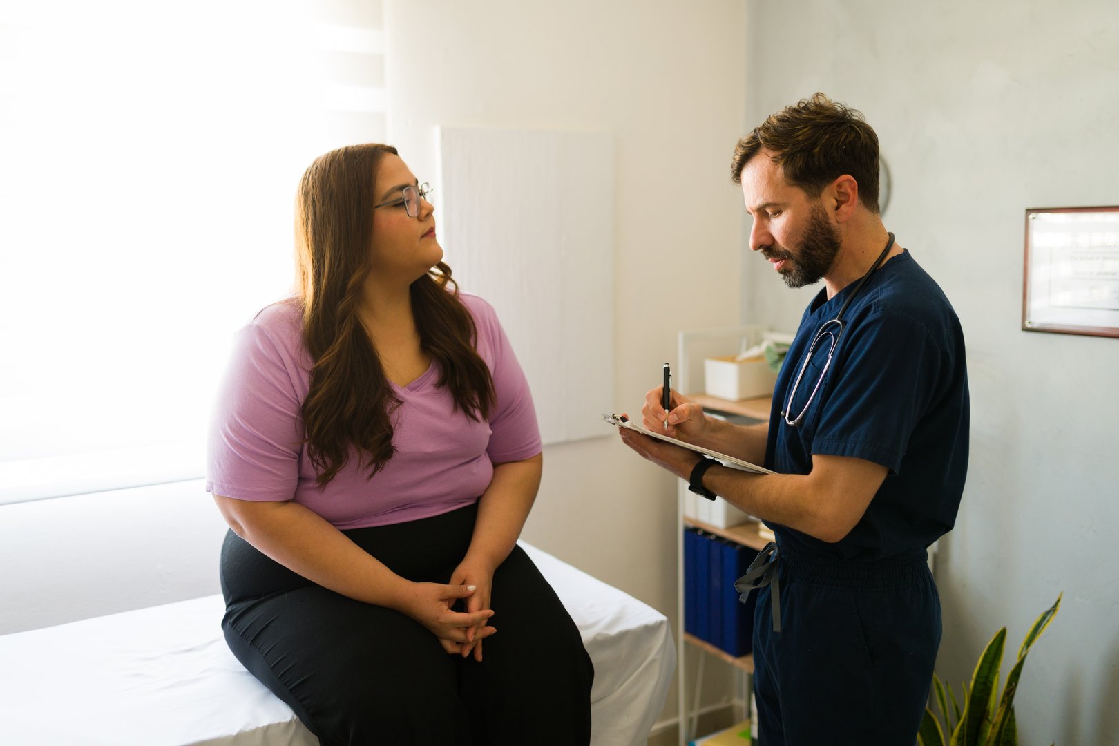 Plus size woman sitting on examination table while talking to doctor taking notes on clipboard during medical consultation in doctor's office