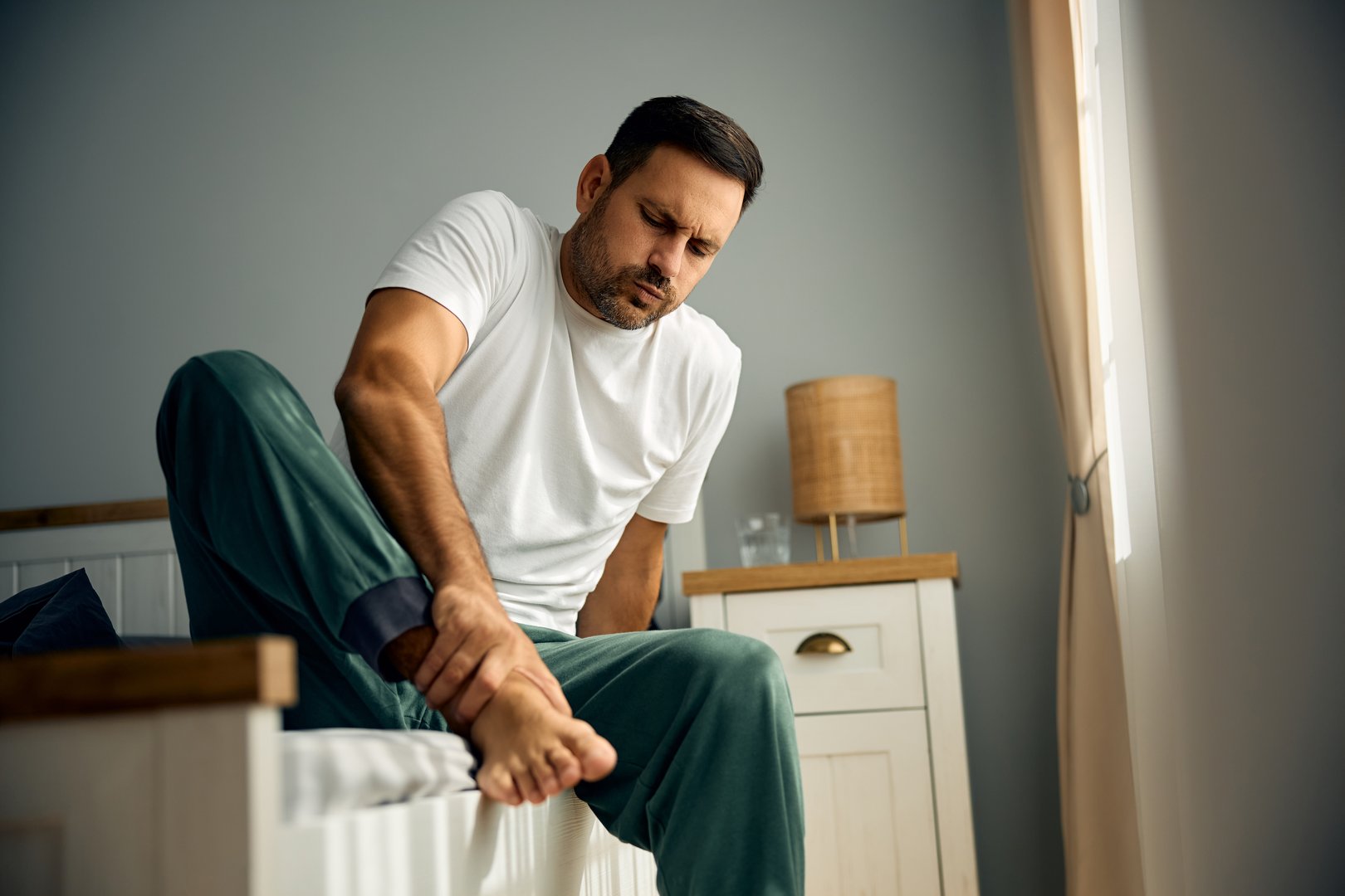 Mid adult man holding his foot in pain in bedroom. Copy space.