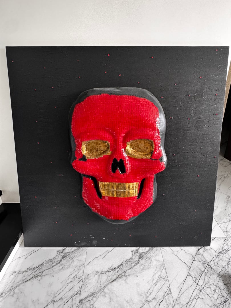 A red skull with gold teeth and eye sockets mounted on a black textured square panel, resting on a marble floor.
