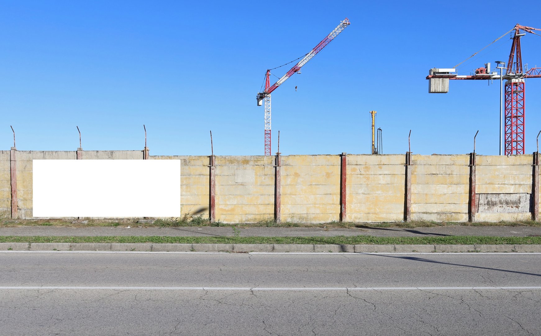 Surrounding wall with tower cranes and building machineries of the construcion site on behind. Blank information board on the left, street in front. Background for copy space.