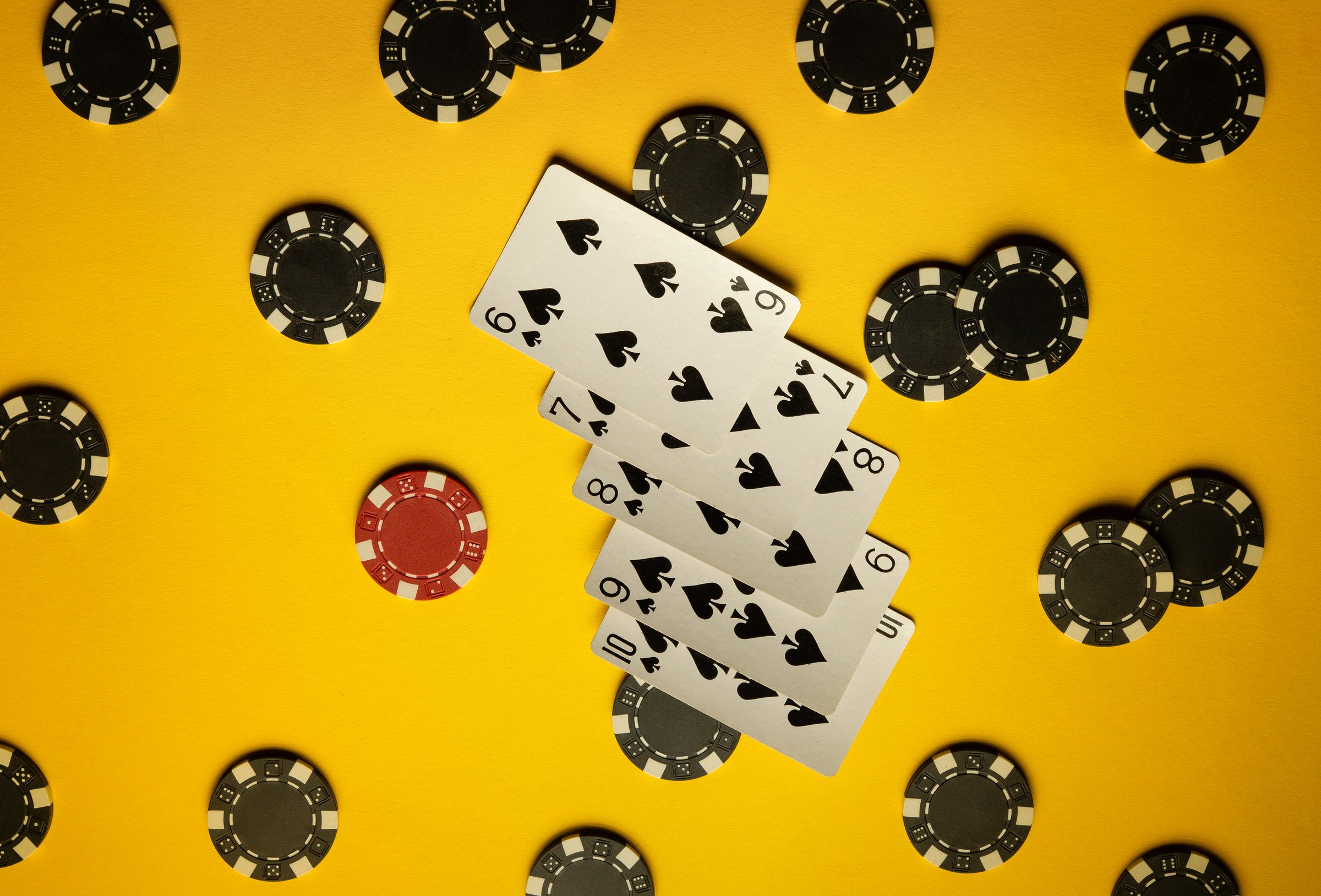 A collection of black poker chips and a red chip scattered around playing cards with spades. The vibrant yellow background adds energy to the gaming setup.