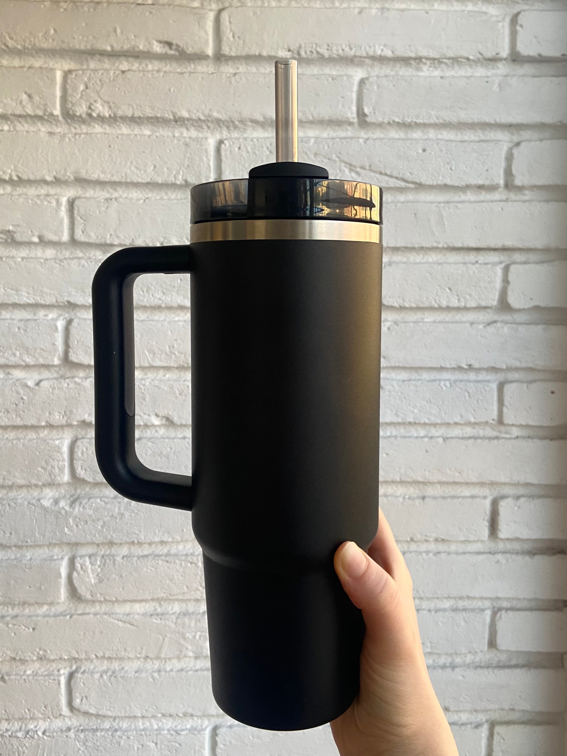 Hand Holding Black Stanley Quencher H2.0 FlowState Tumbler With Handle Against White Brick Wall. High quality photo