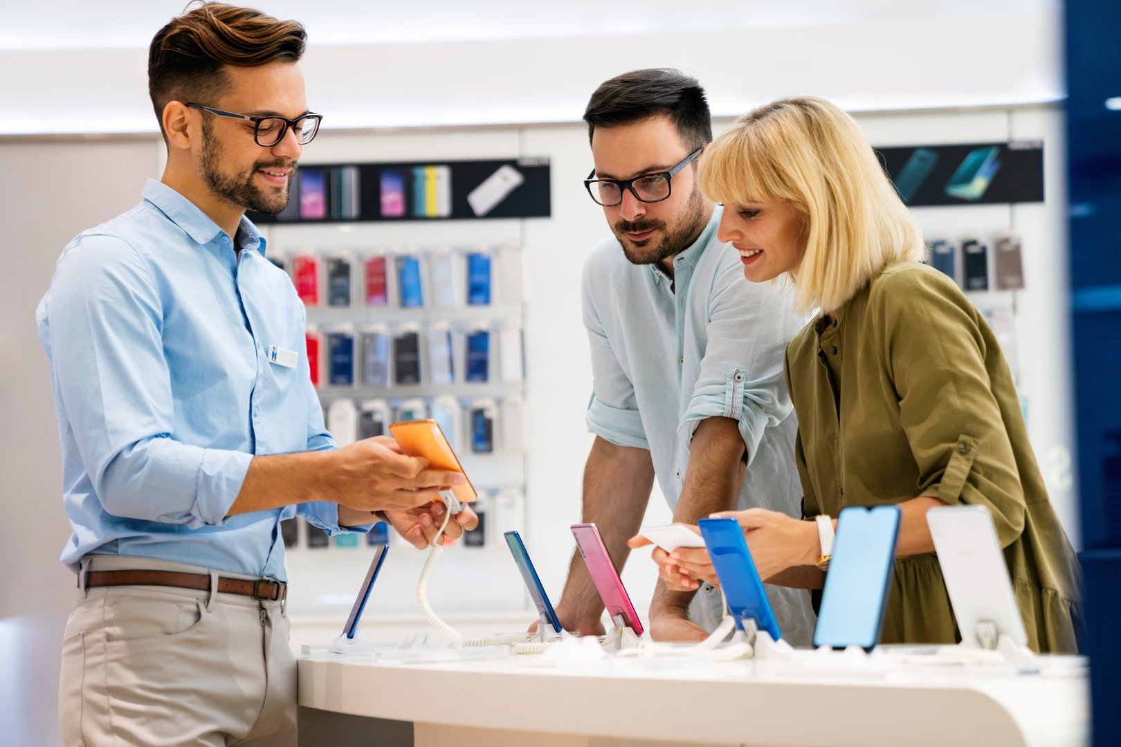 Happy seller man helping to a couple to buy a new digital smart device in tech store. Technology people concept