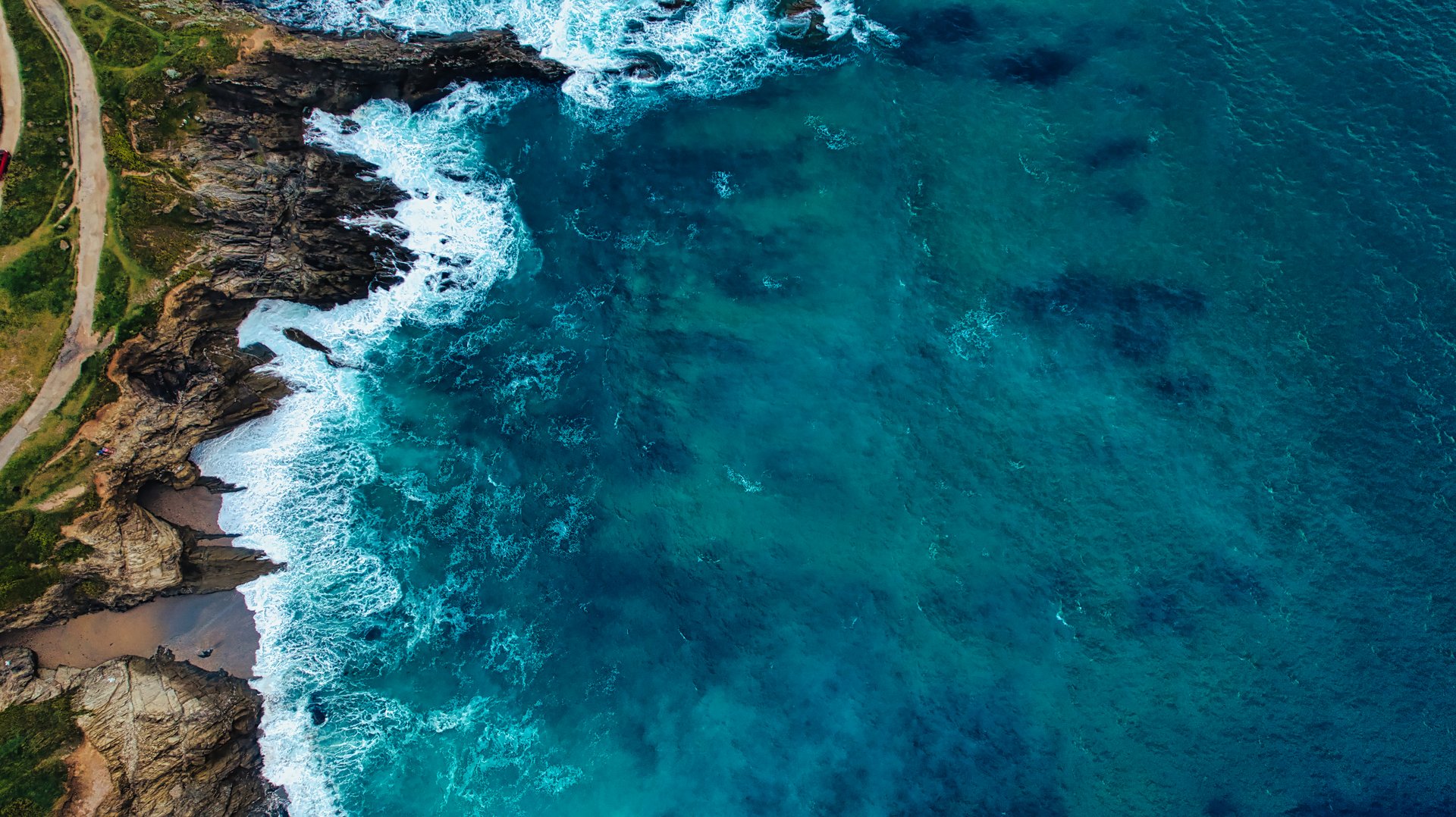Aerial view of a rocky coastline meeting the ocean, with waves crashing against the shore. The water is a vibrant blue, showcasing different shades and textures. The land features a winding path along the cliffs.