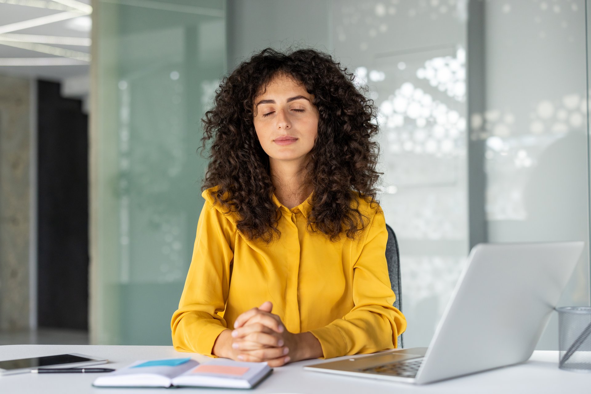 Businesswoman embracing tranquility at a desk, eyes closed, practicing meditation for mindfulness and stress relief. Laptop and notepad symbolize modern office setting and work-life balance.