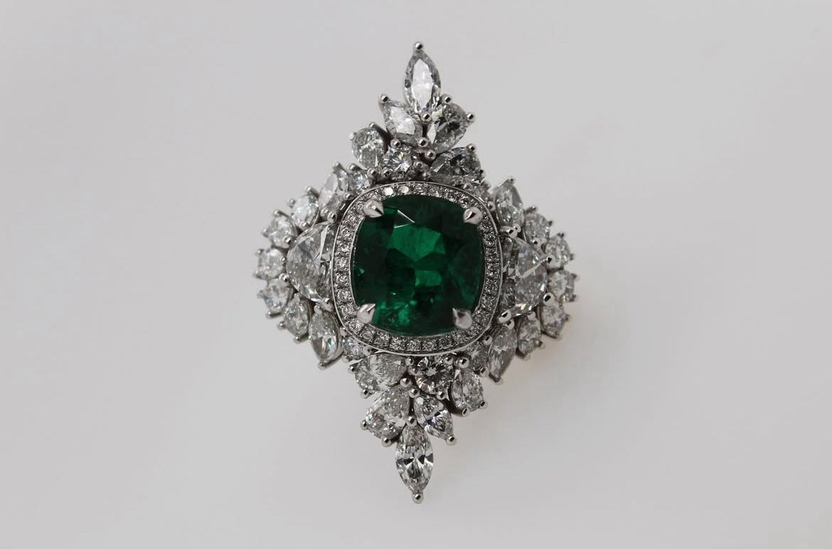 Intricate emerald and diamond ring with a central green gemstone surrounded by a detailed diamond arrangement.