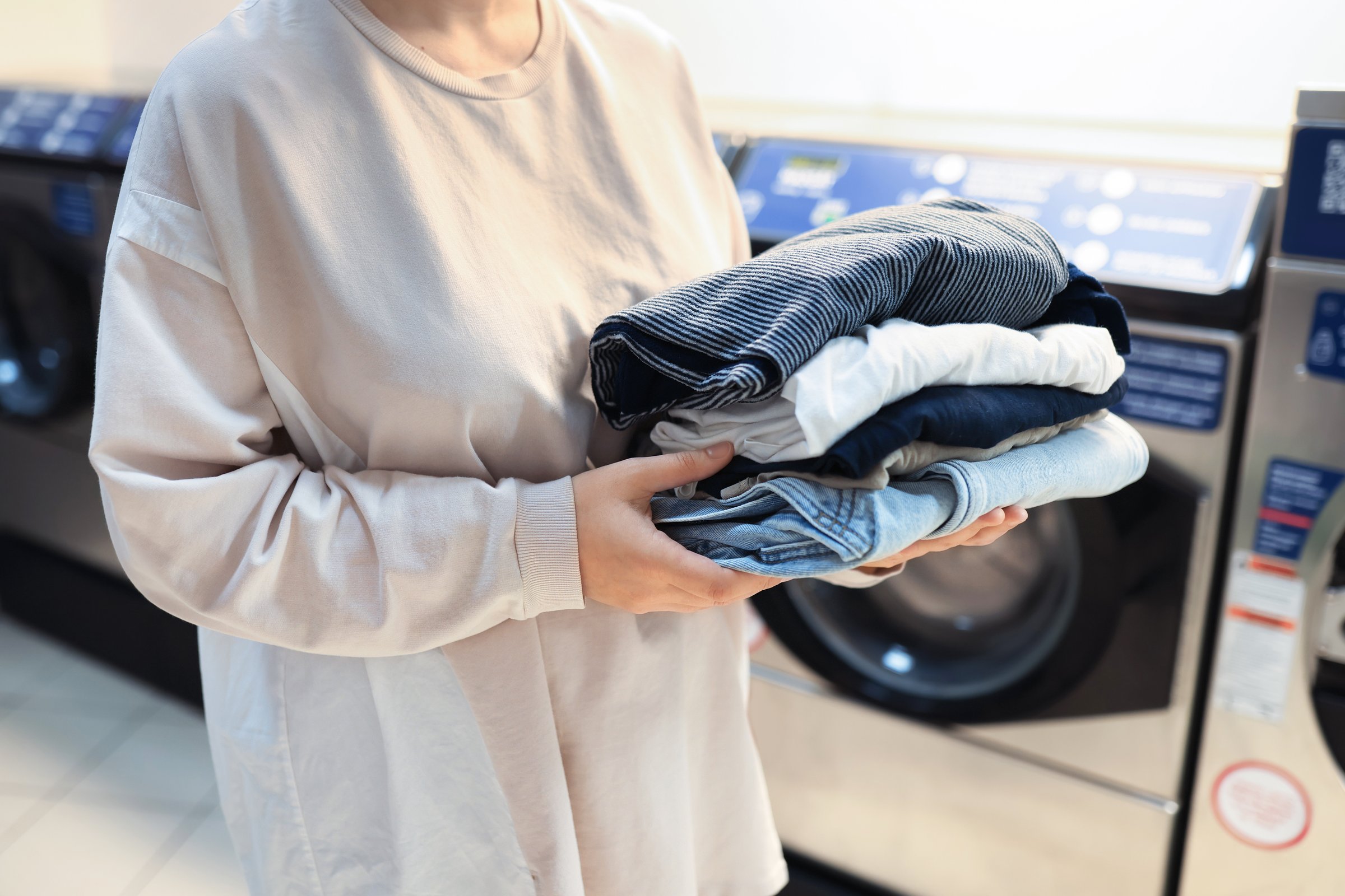 Woman holding stack of folded clean clothes at public laundromat with washing machines in background. High quality photo