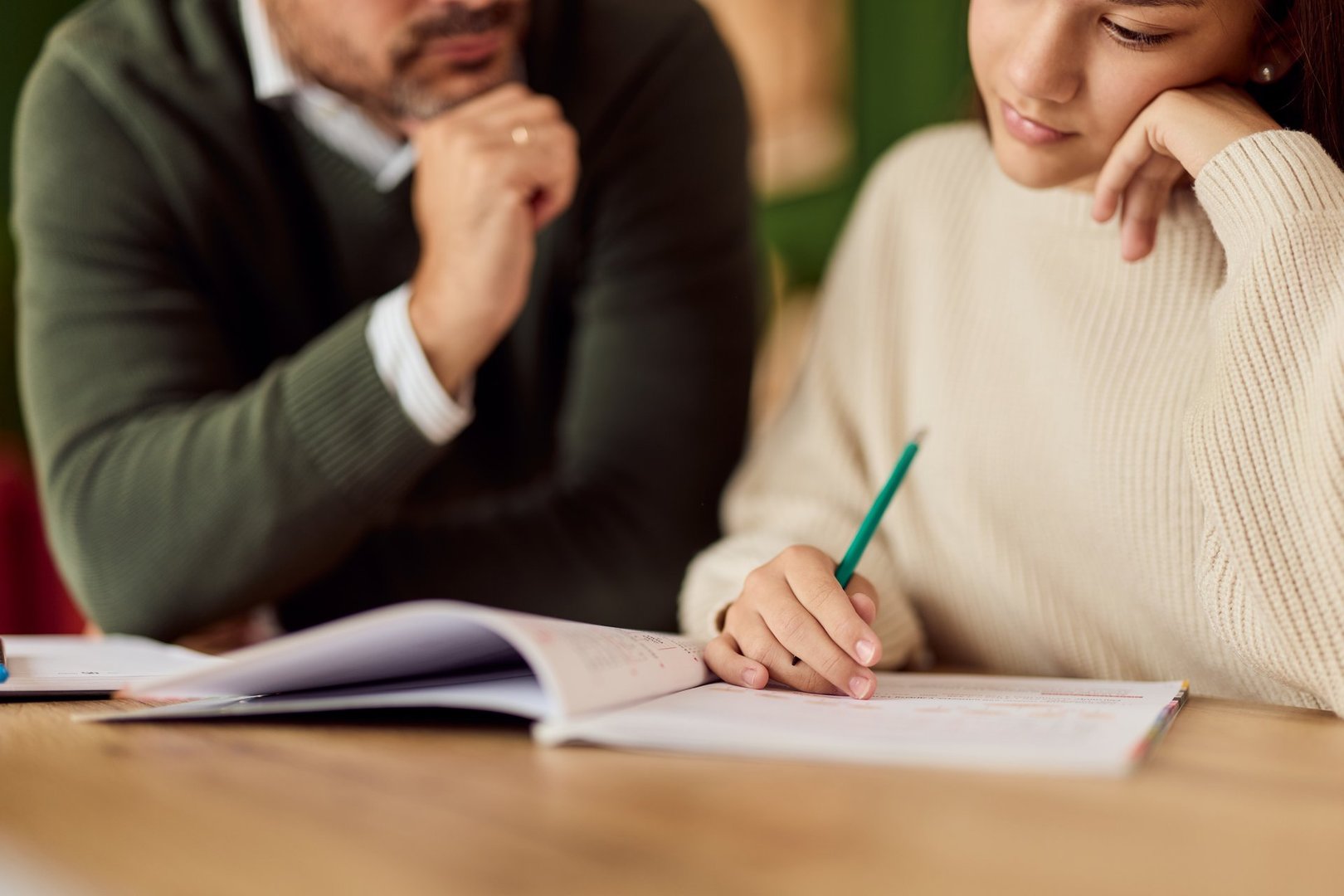 A man and a young girl focus on homework at a wooden desk, writing and reviewing notes