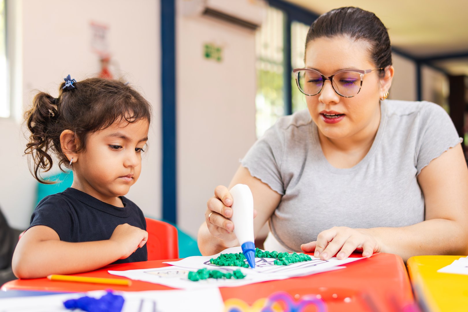 Teacher helping little girl gluing colorful pieces of paper on a drawing in a preschool classroom