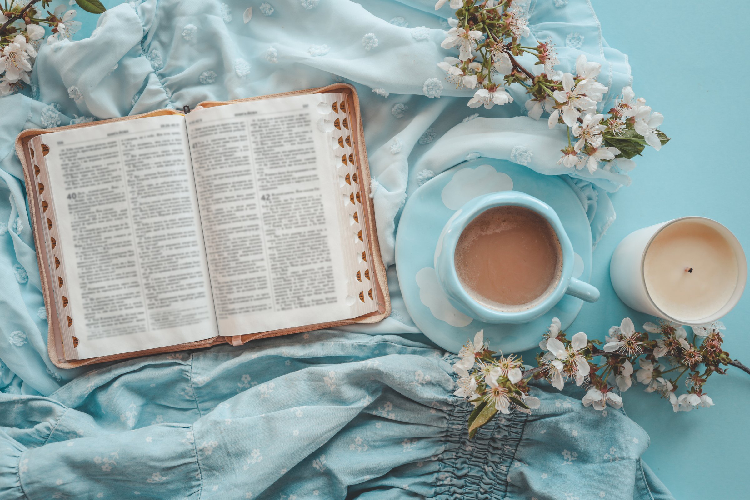 Open Bible with a cup of coffee on a blue saucer, surrounded by delicate white spring blossoms and soft blue fabric, creating a peaceful and spiritual atmosphere.