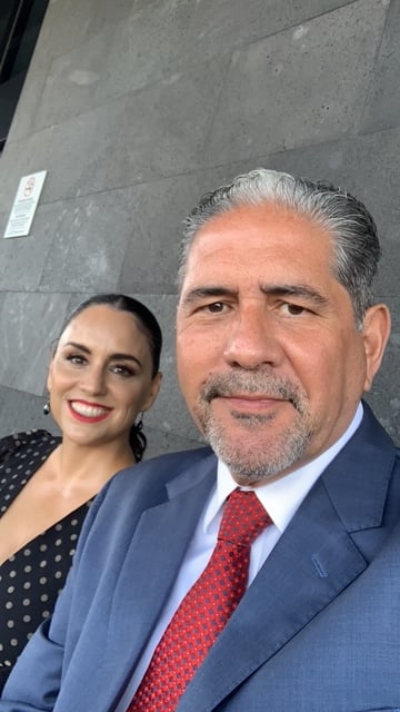 A man in a blue suit and red tie and a woman in a black polka dot dress smiling while seated against a gray stone wall.
