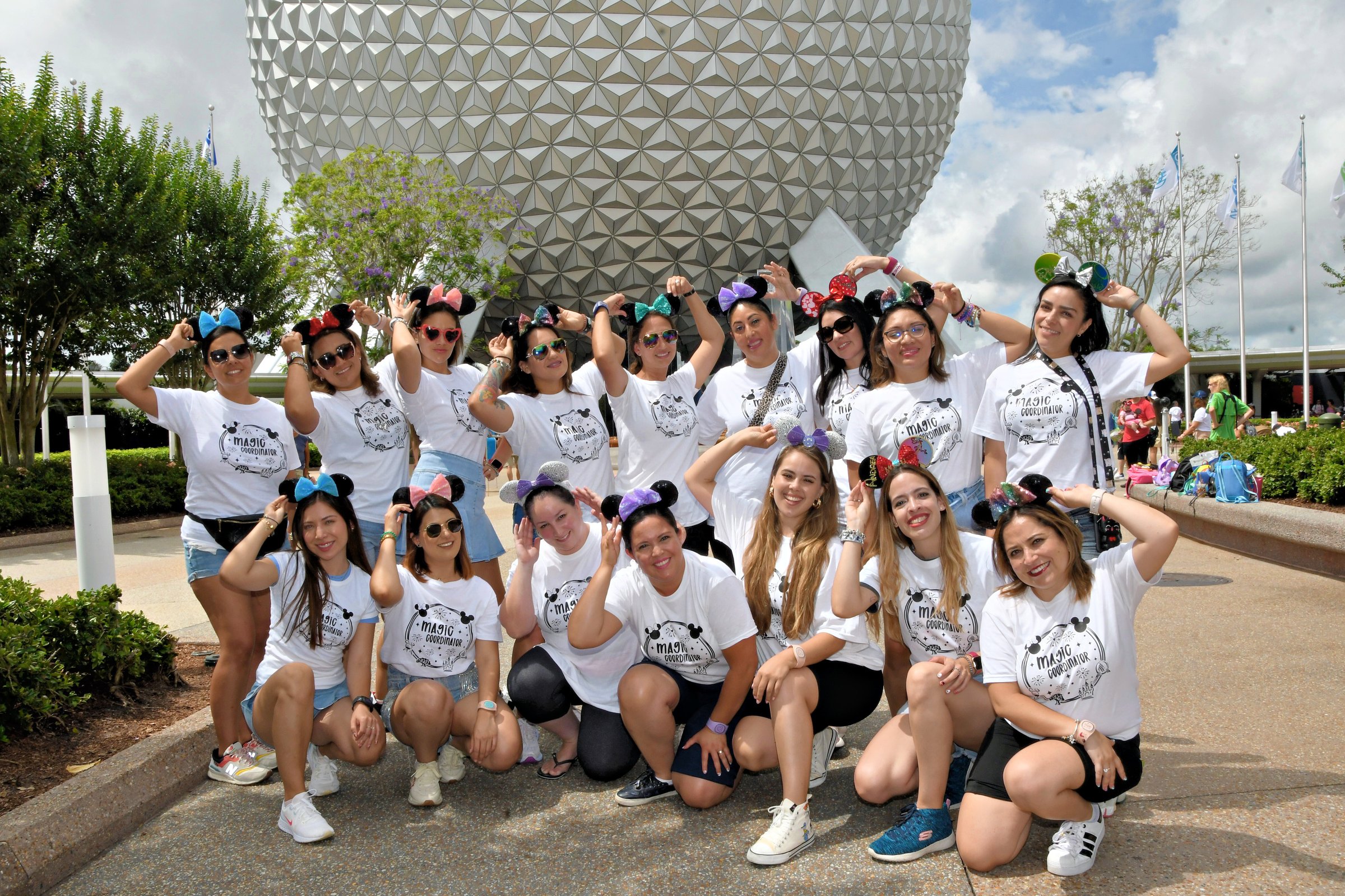 Group of women in matching shirts and Mickey ears pose in front of a large geodesic sphere at an amusement park.