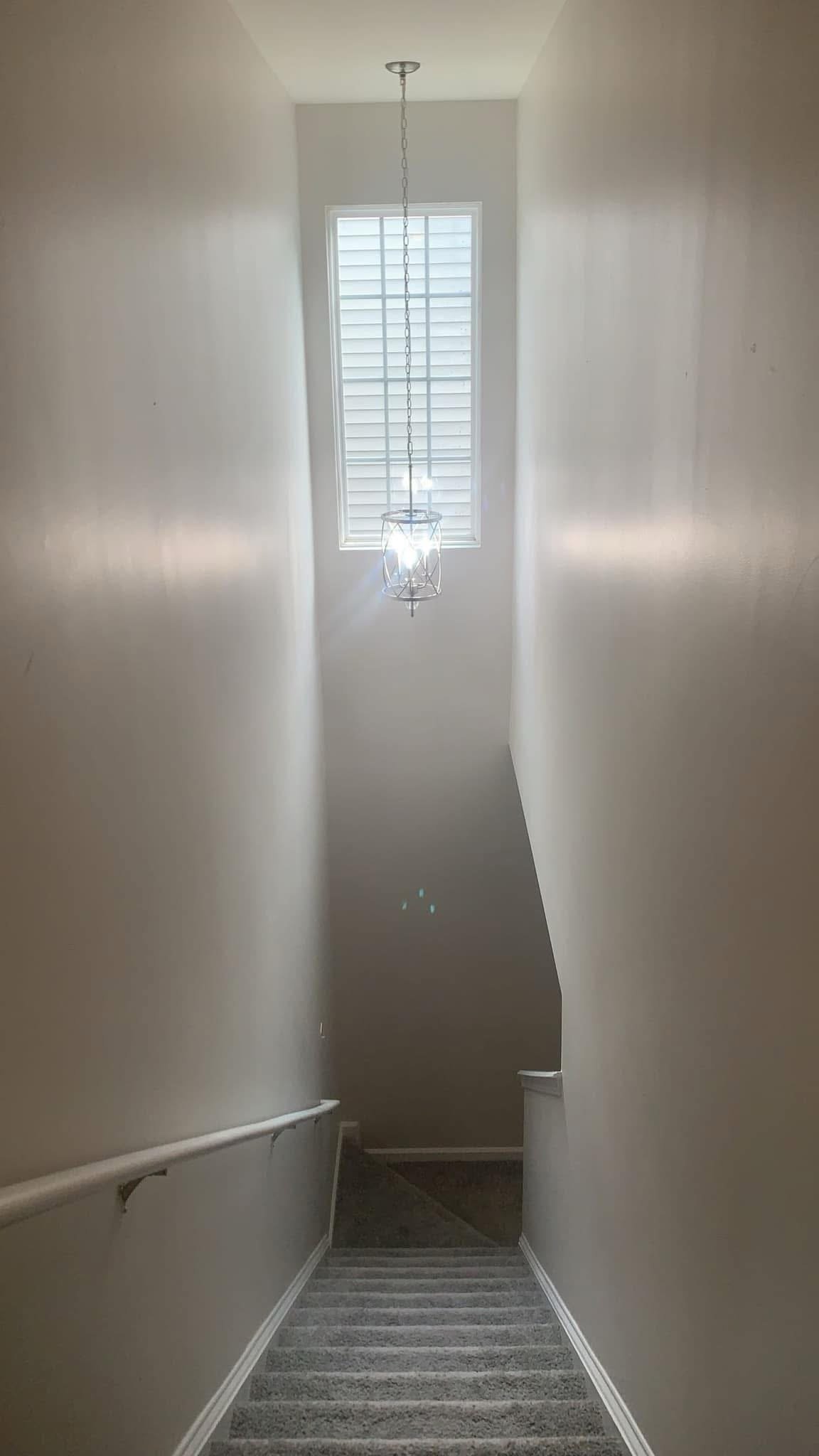 A staircase with gray carpet leads downward, illuminated by a hanging light fixture and a window with blinds above.