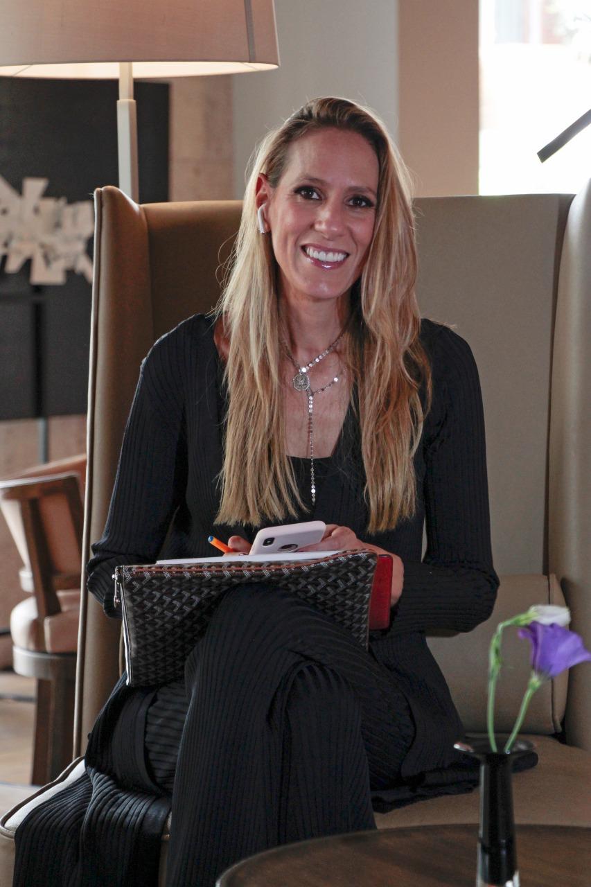 A woman with long blonde hair smiles while sitting in a chair, holding a notebook and phone, with a purple flower in the foreground.