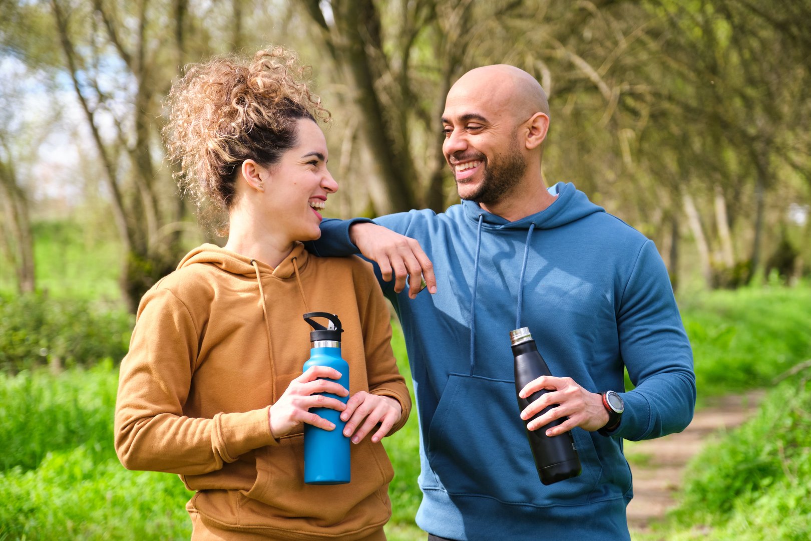 A man and a woman are smiling and holding water bottles. The man is wearing a blue hoodie and the woman is wearing a brown hoodie. They are exercising together in a park.