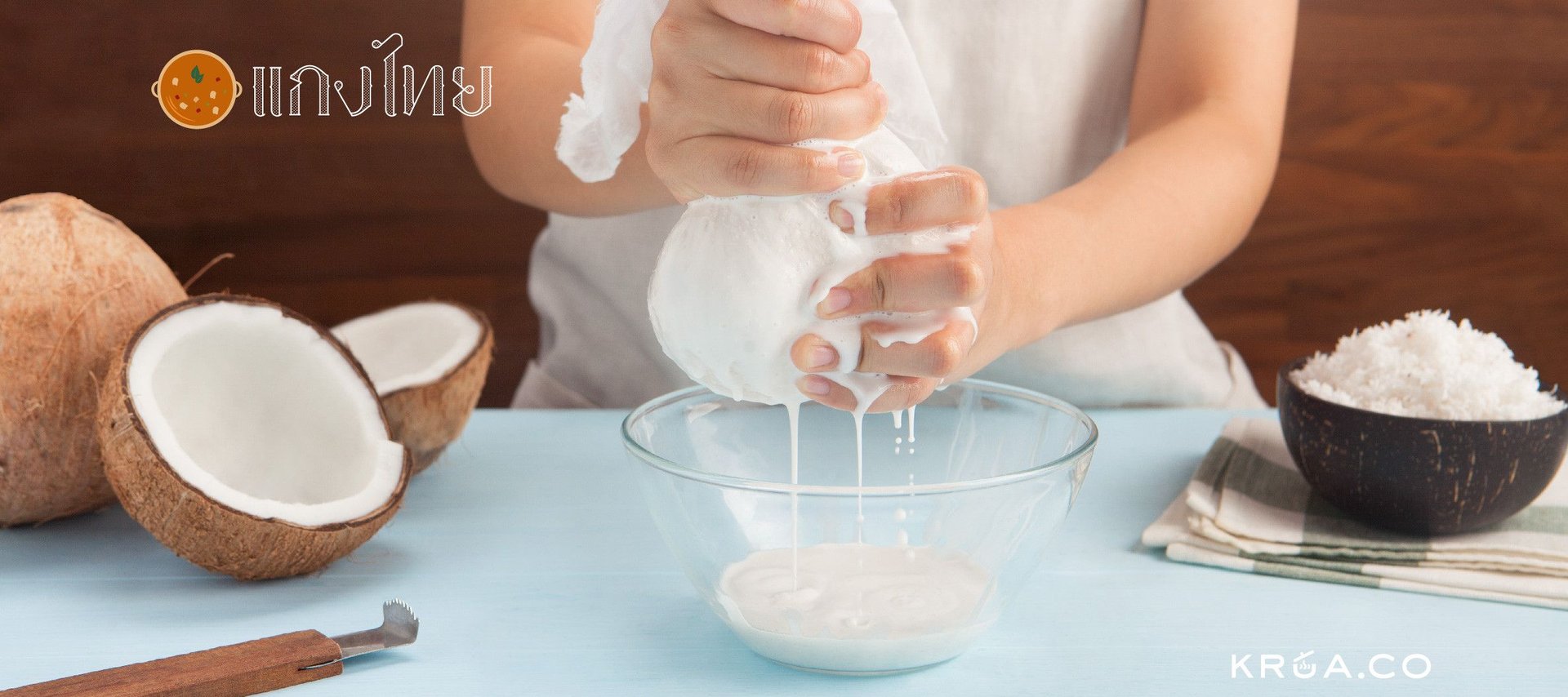 Person squeezing coconut milk into a bowl, surrounded by coconut halves and grated coconut on a table.