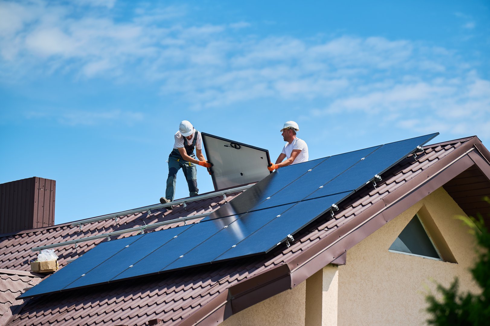 Workers building solar panel system on roof of house. Two men installers in helmets carrying photovoltaic solar module outdoors. Alternative, green and renewable energy generation concept.