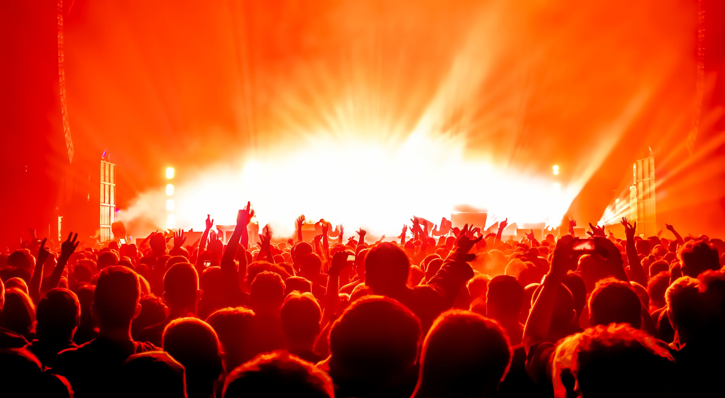 Concert crowd with raised hands against a bright, vibrant orange-lit stage in the background.