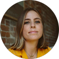 A person with shoulder-length hair wearing a yellow top, standing against a brick wall backdrop.