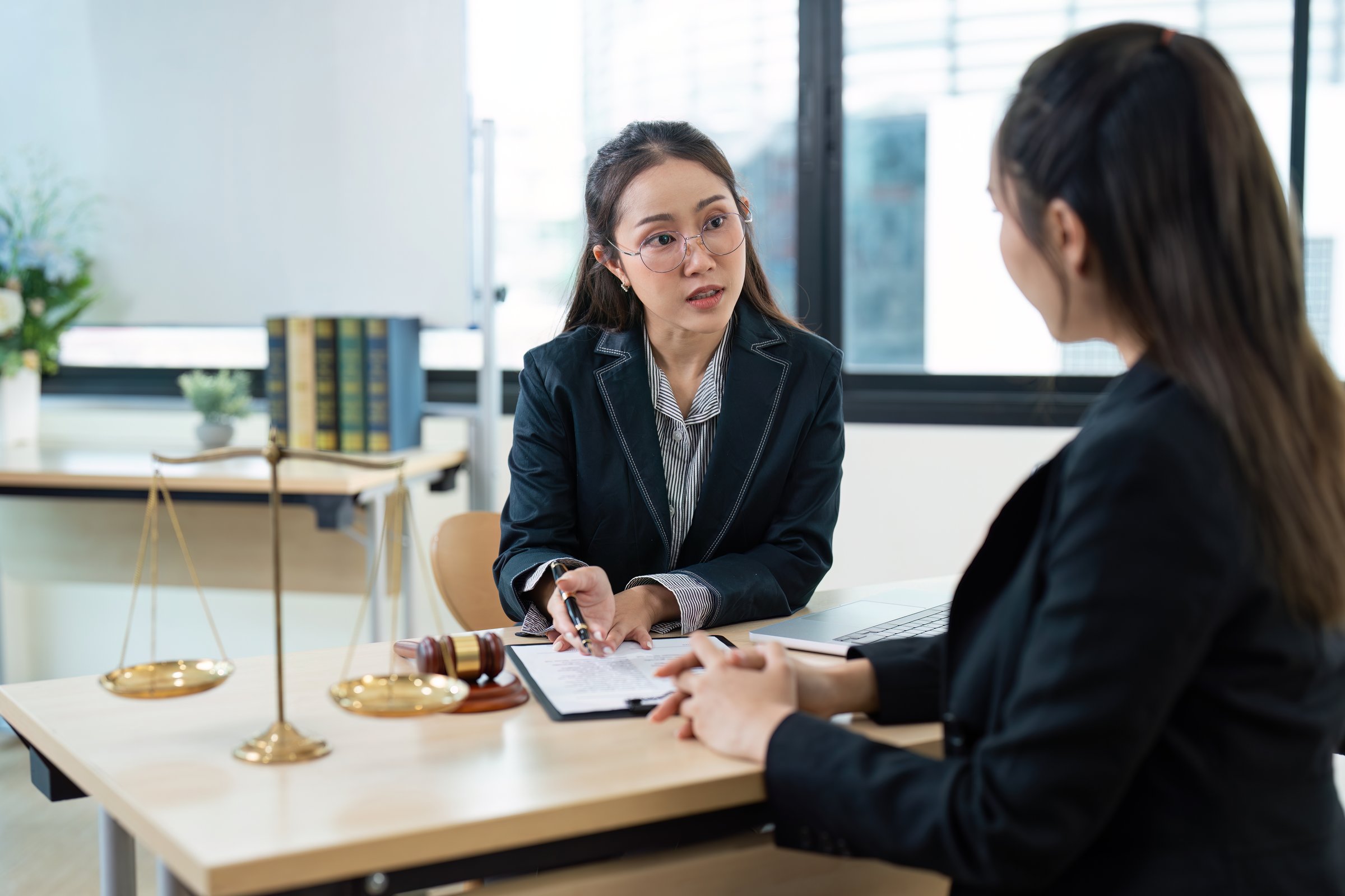 A professional female lawyer consulting a client in a modern office setting, discussing legal documents with scales of justice on the desk.