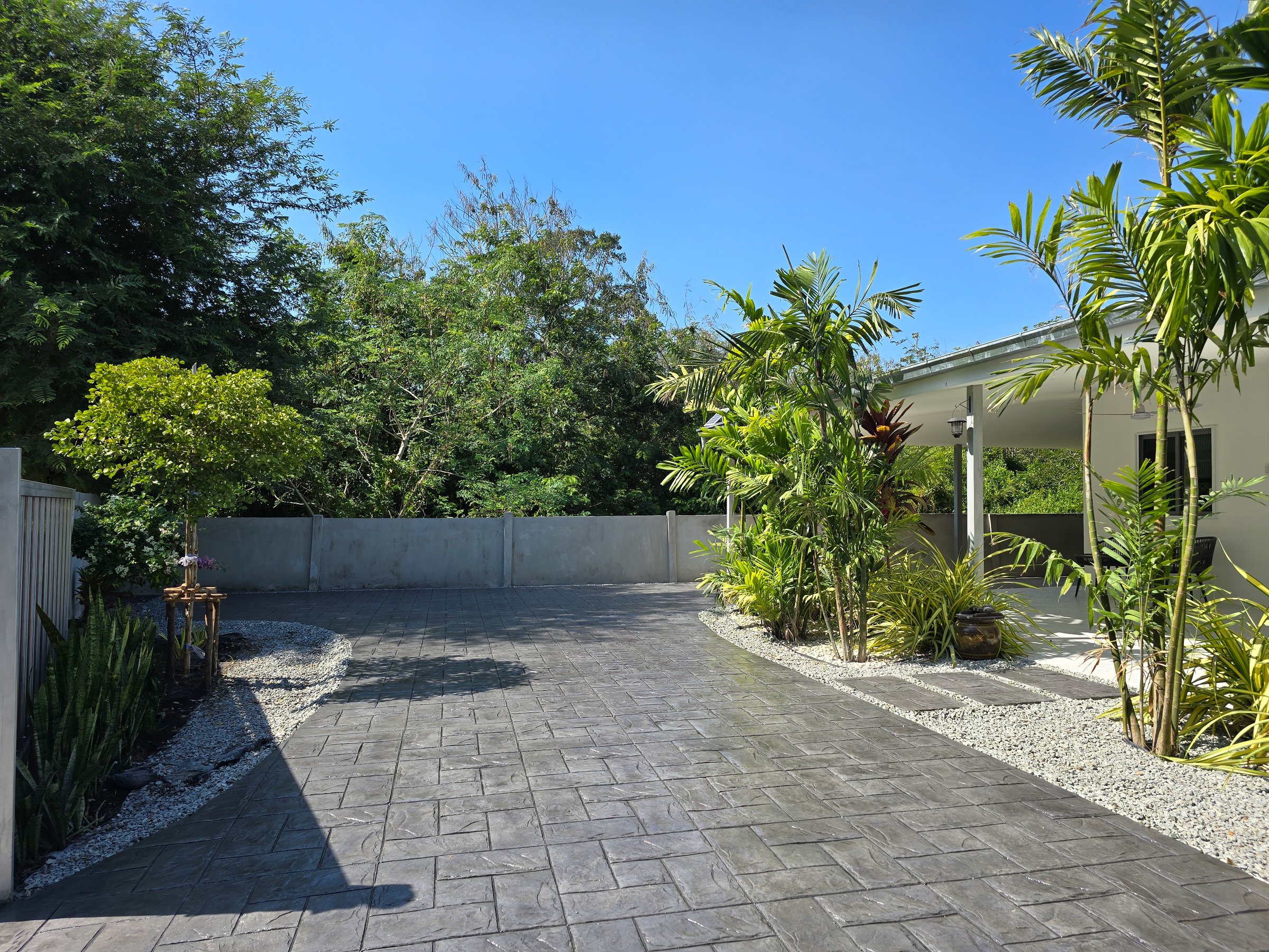 In Pattaya, Thailand, a stunning stamped concrete driveway showcases intricate designs. Lush plants and trees frame this serene outdoor space, a Thai Suburban area with a modern family house