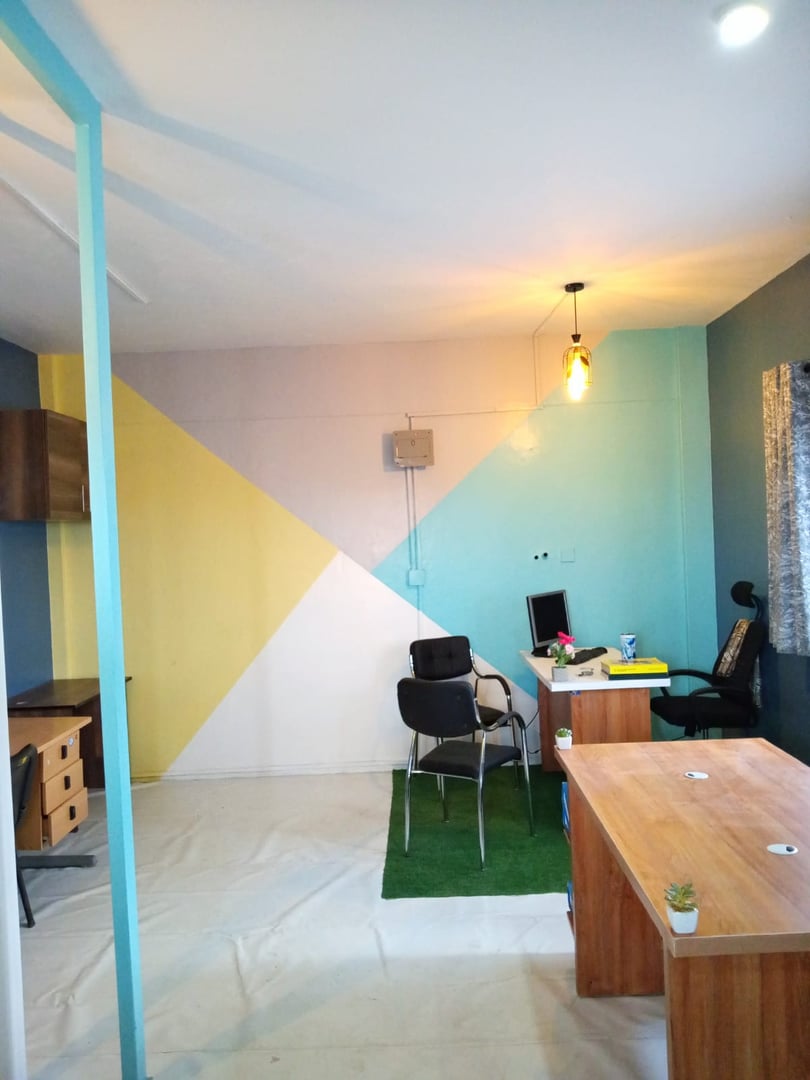 Office space with colorful geometric wall, desks, chairs, and a hanging light bulb. Contains plants and a computer.