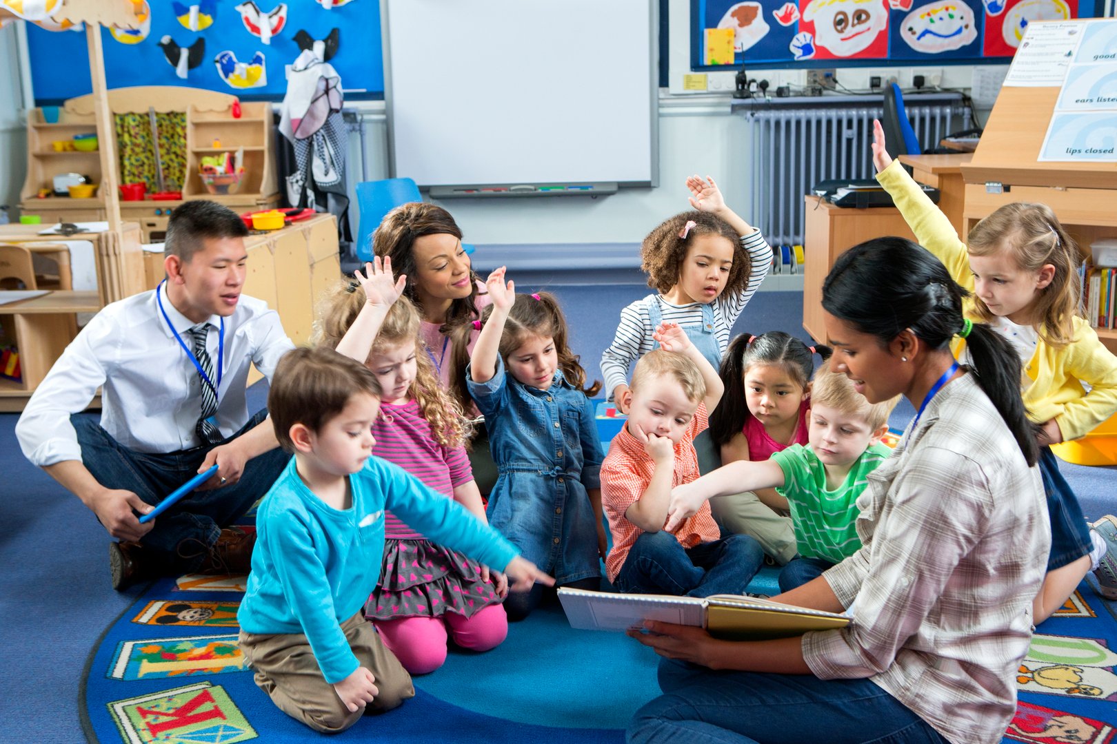 Female teacher giving a lesson to nursery students. They are sitting on the floor and some have their hands up to ask a question.