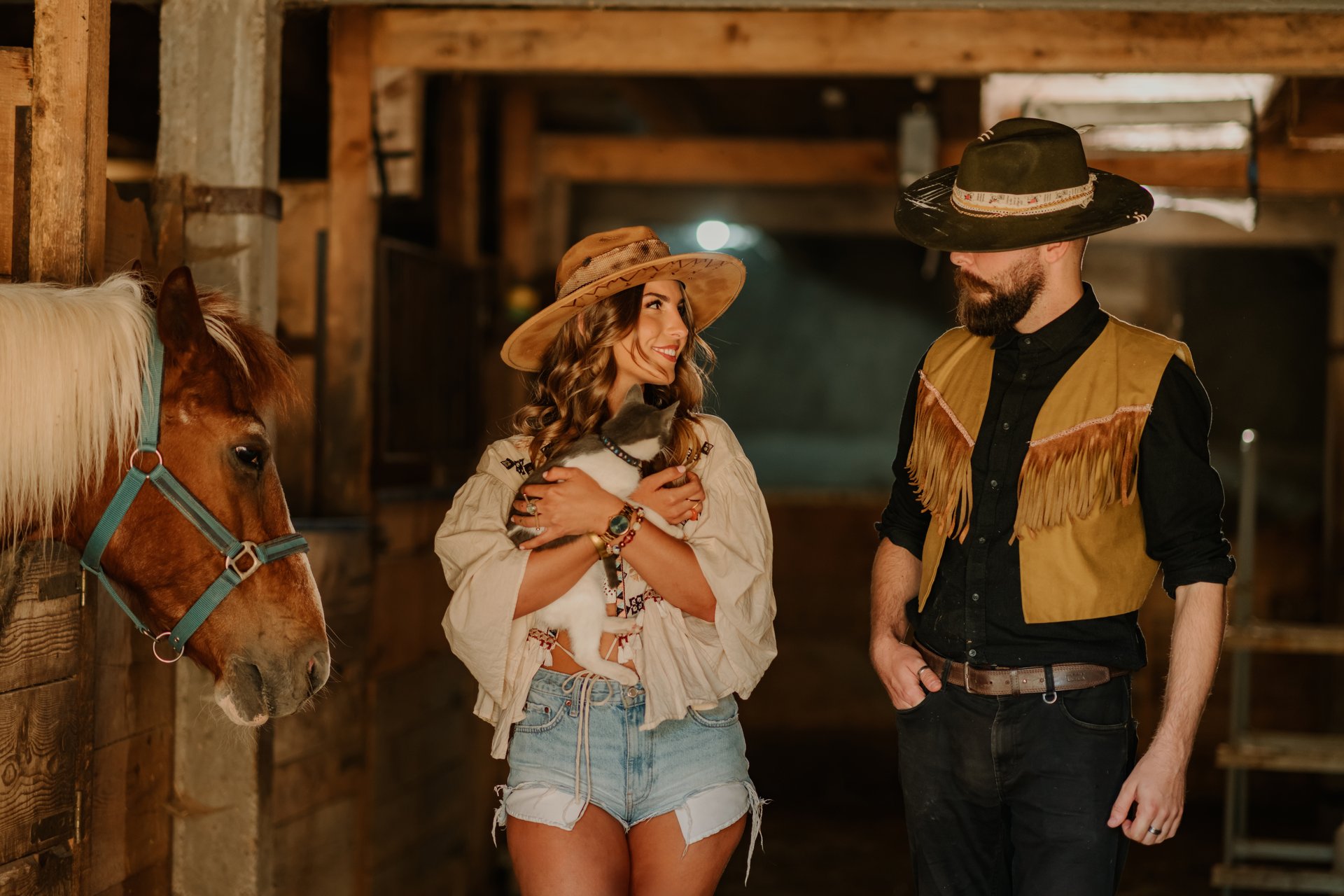 Portrait of young affectionate rancher couple in western styled outfit with domestic cat in hands walking around horse stable at ranch and smiling at each other. Animal lovers taking care of animals.