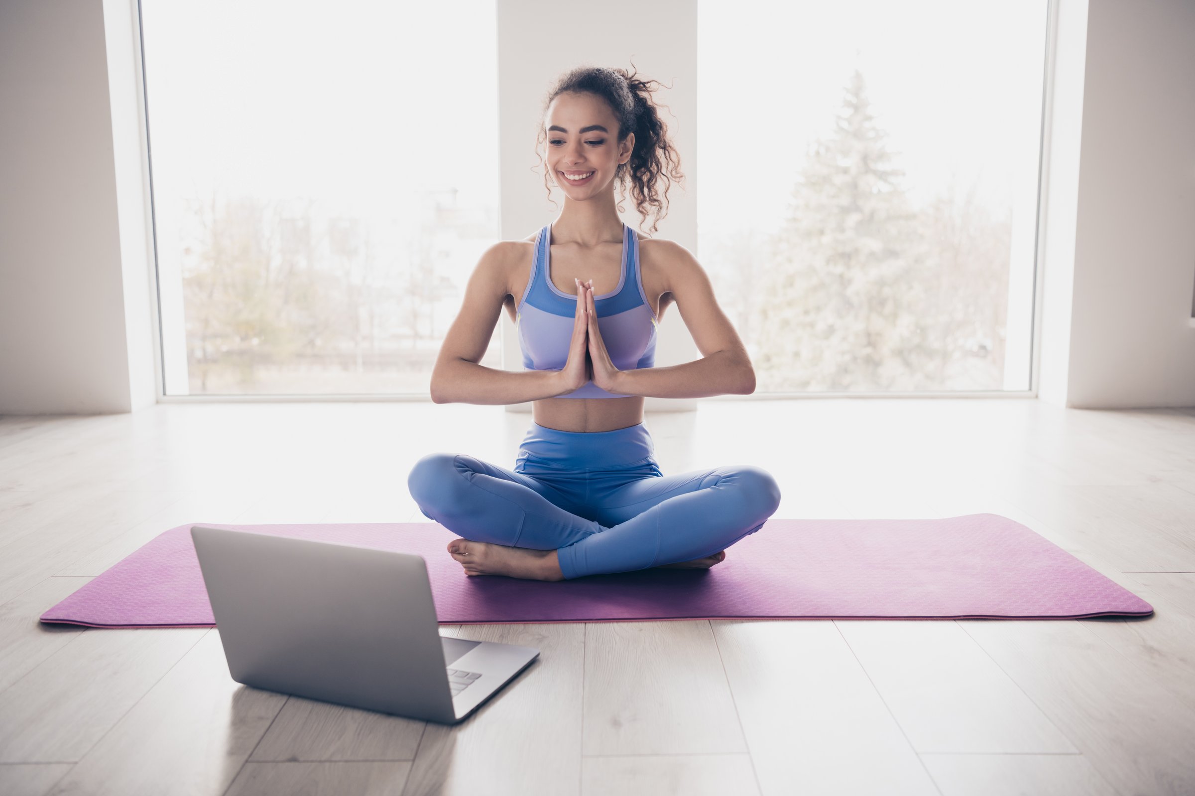 A young woman enjoys a yoga session at home, following instructions on a laptop. She is seated on a yoga mat in a bright and modern interior.