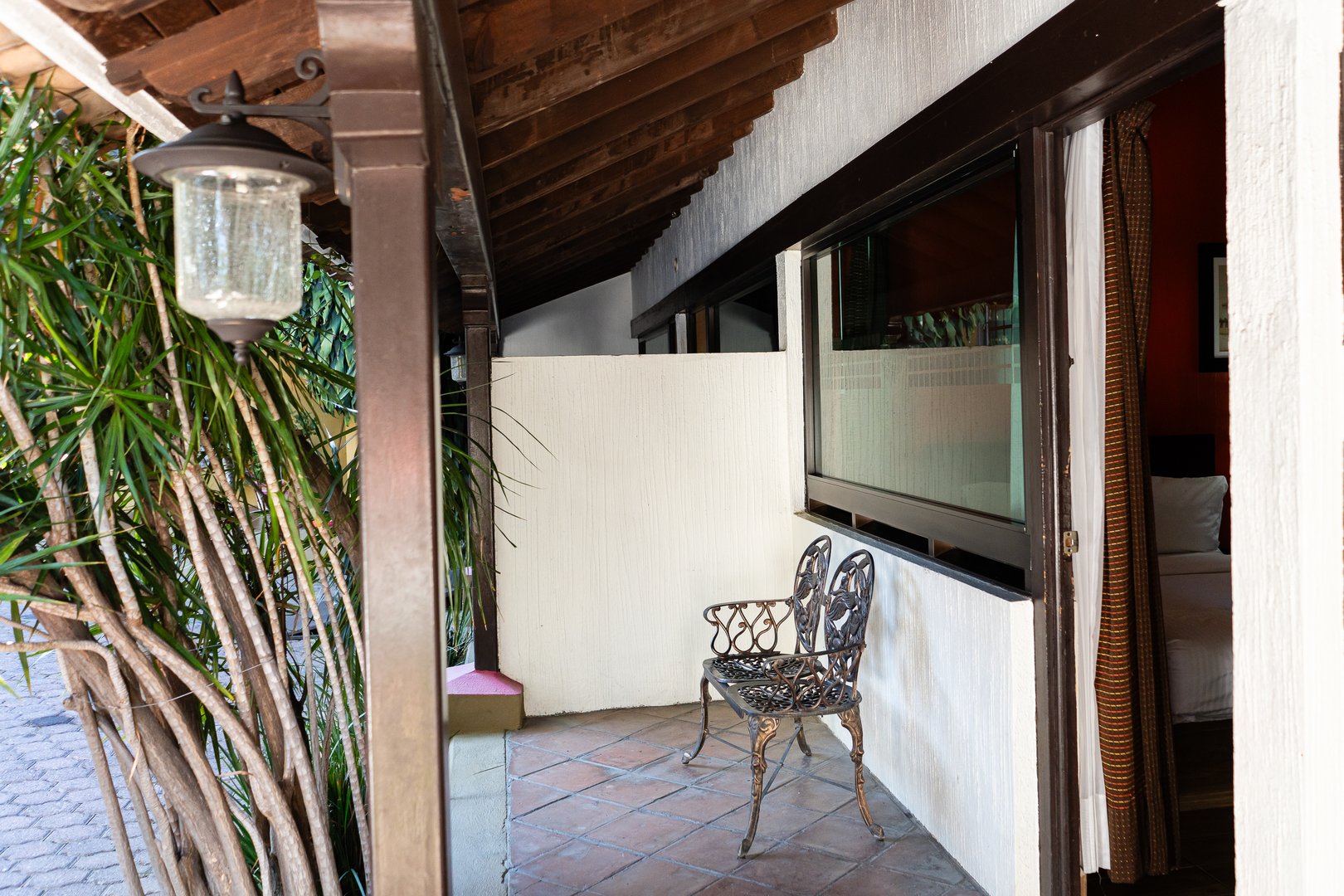 Outdoor patio with decorative metal chair, tiled floor, and partial view of a bedroom through a window.
