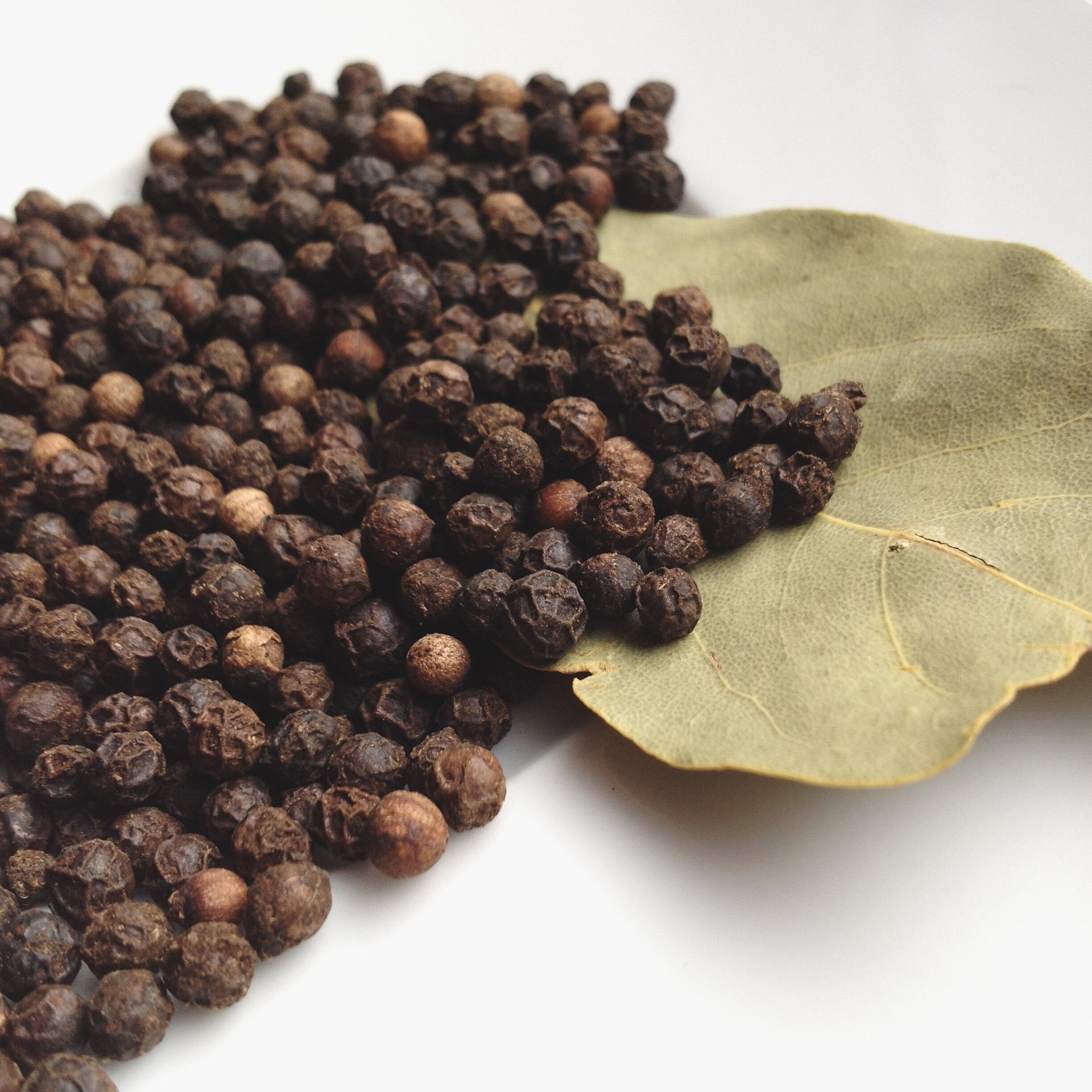 Black peppercorns scattered beside two bay leaves on a white surface.