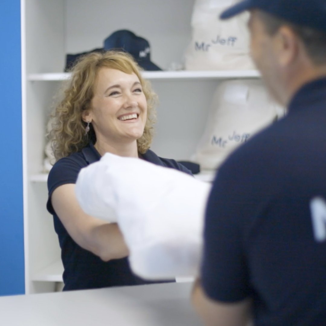 Woman smiling while handing a folded garment to a uniformed worker at a laundry service.